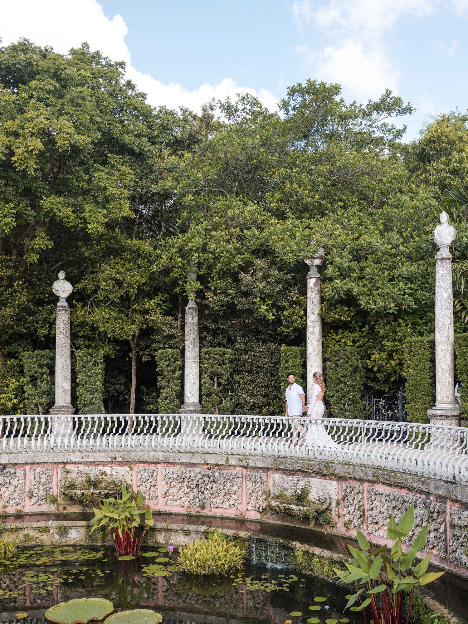 Bride and groom walk hand in hand along an elegant curved stone terrace surrounded by lush tropical gardens and classical marble ccolumns.