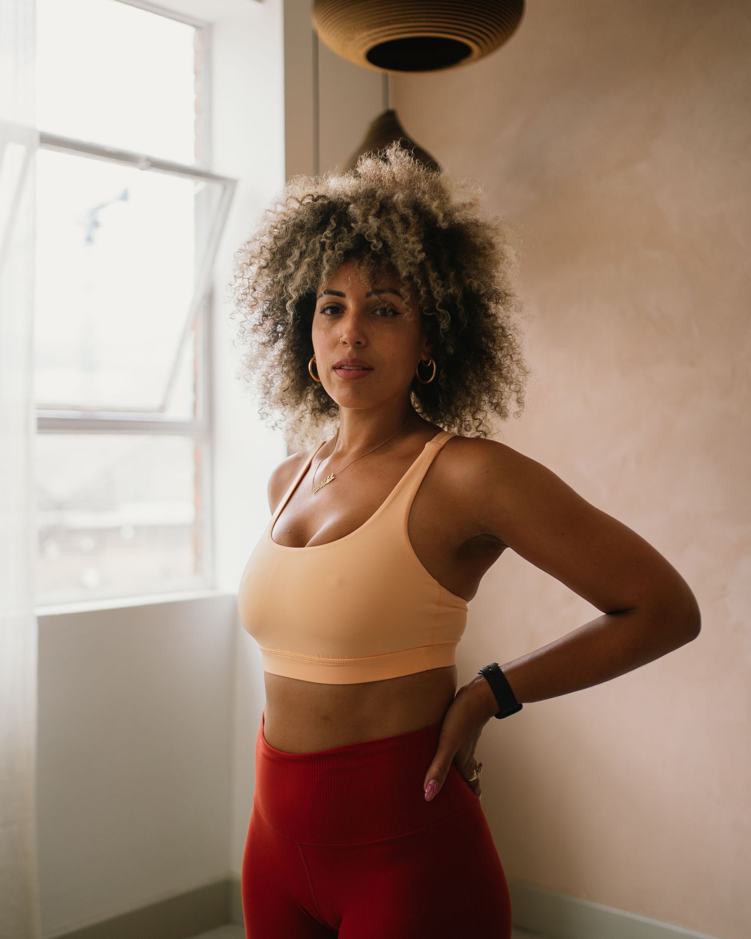 Woman in beige sports bra and red leggings poses confidently indoors near a window with natural light.