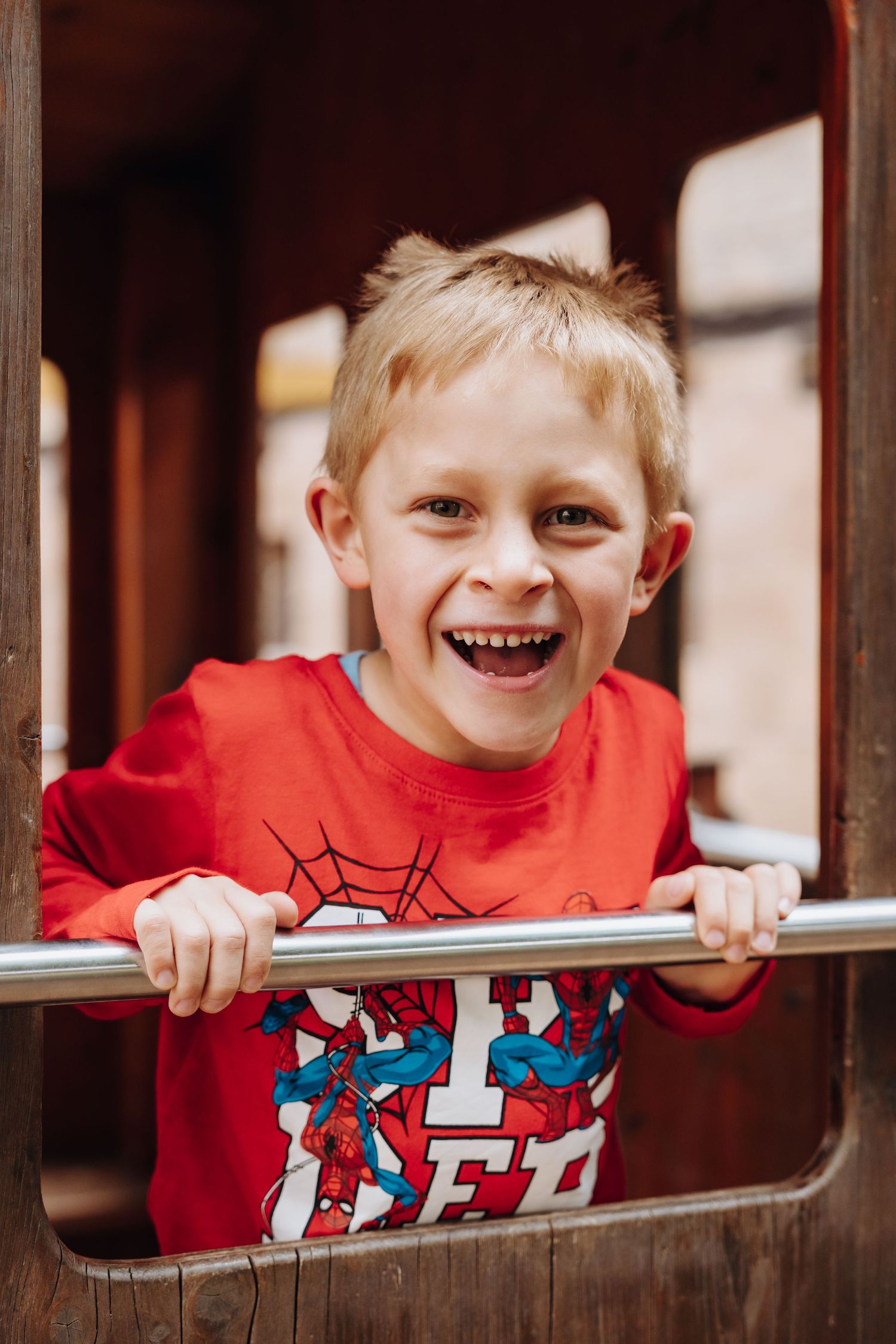 Smiling blonde boy in red Spider-Man shirt playing on wooden playground equipment, gripping a metal bar.