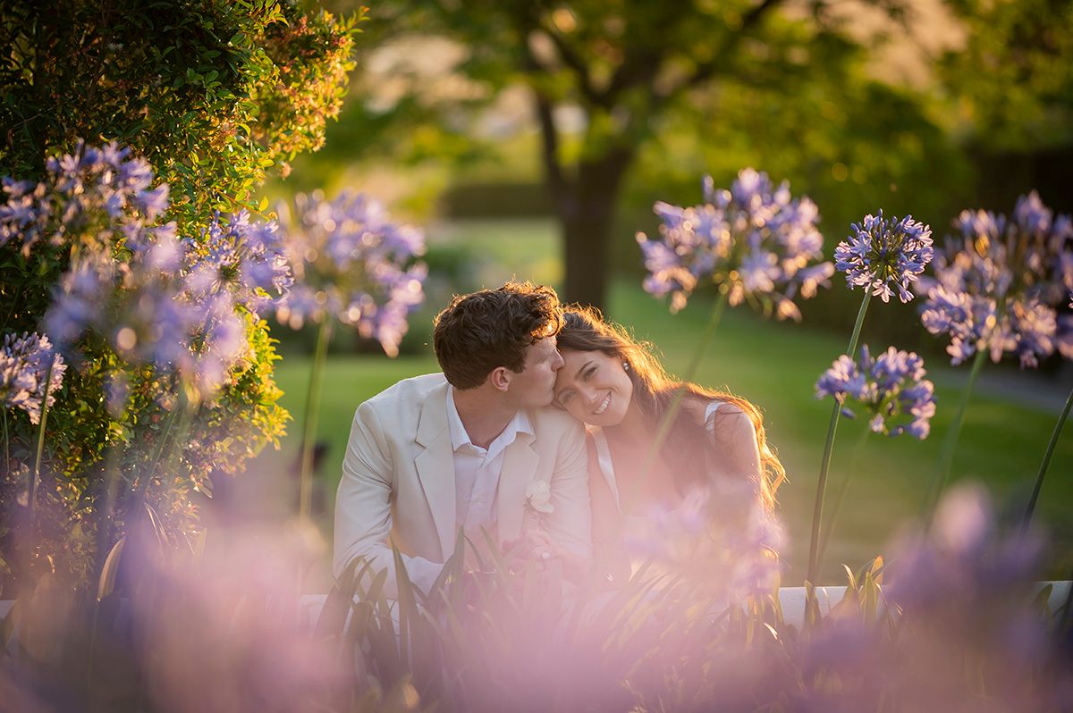 A couple embraces in a romantic garden setting with soft sunlight filtering through pink cherry blossoms.