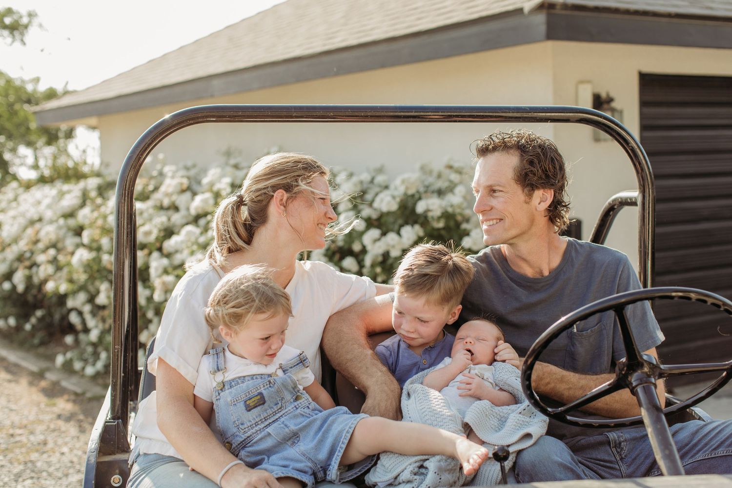 Happy family with three young children sitting together on a vintage vehicle outdoors near a white farmhouse.