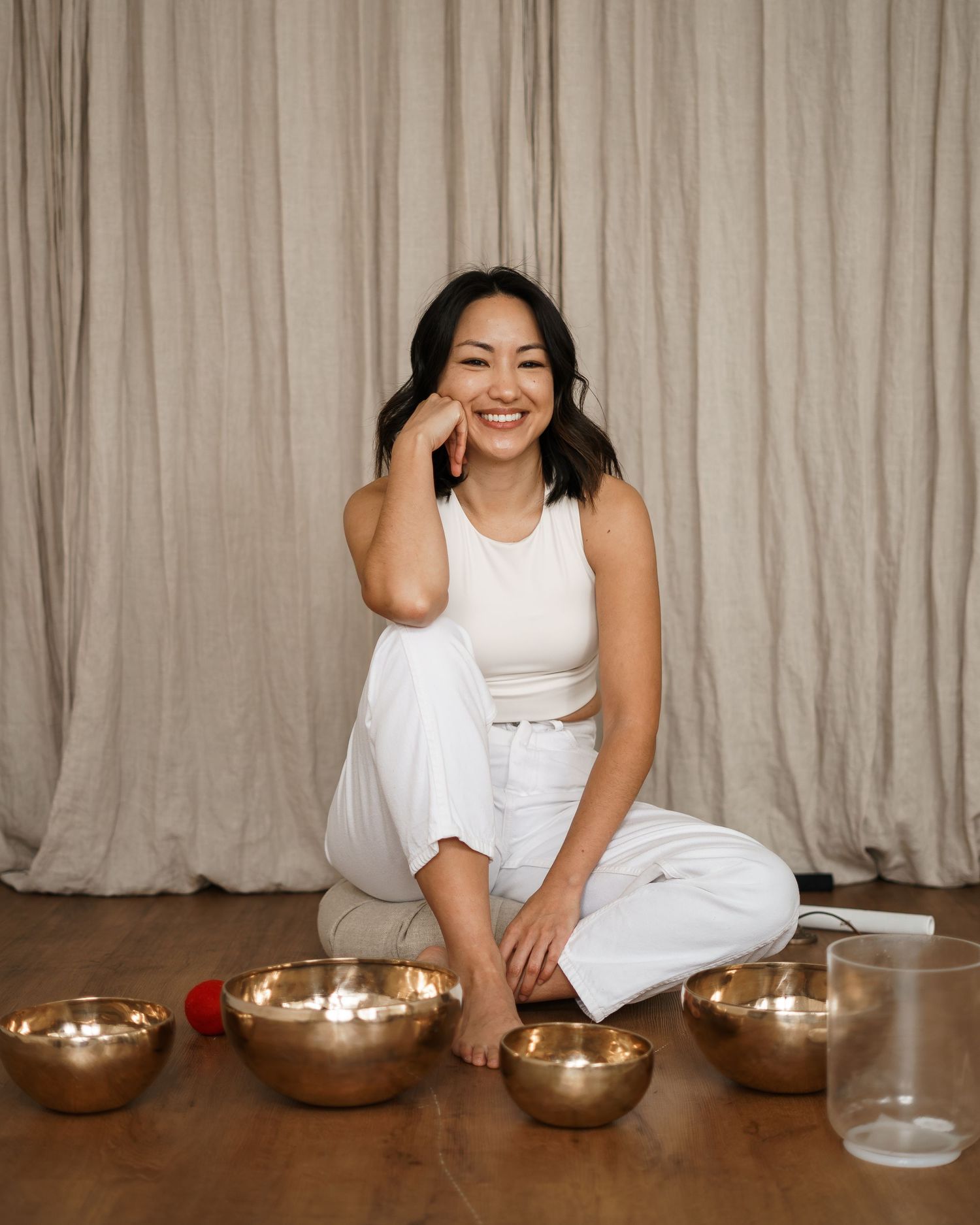 Smiling woman in white outfit sitting on floor with Tibetan singing bowls and crystal bowl.