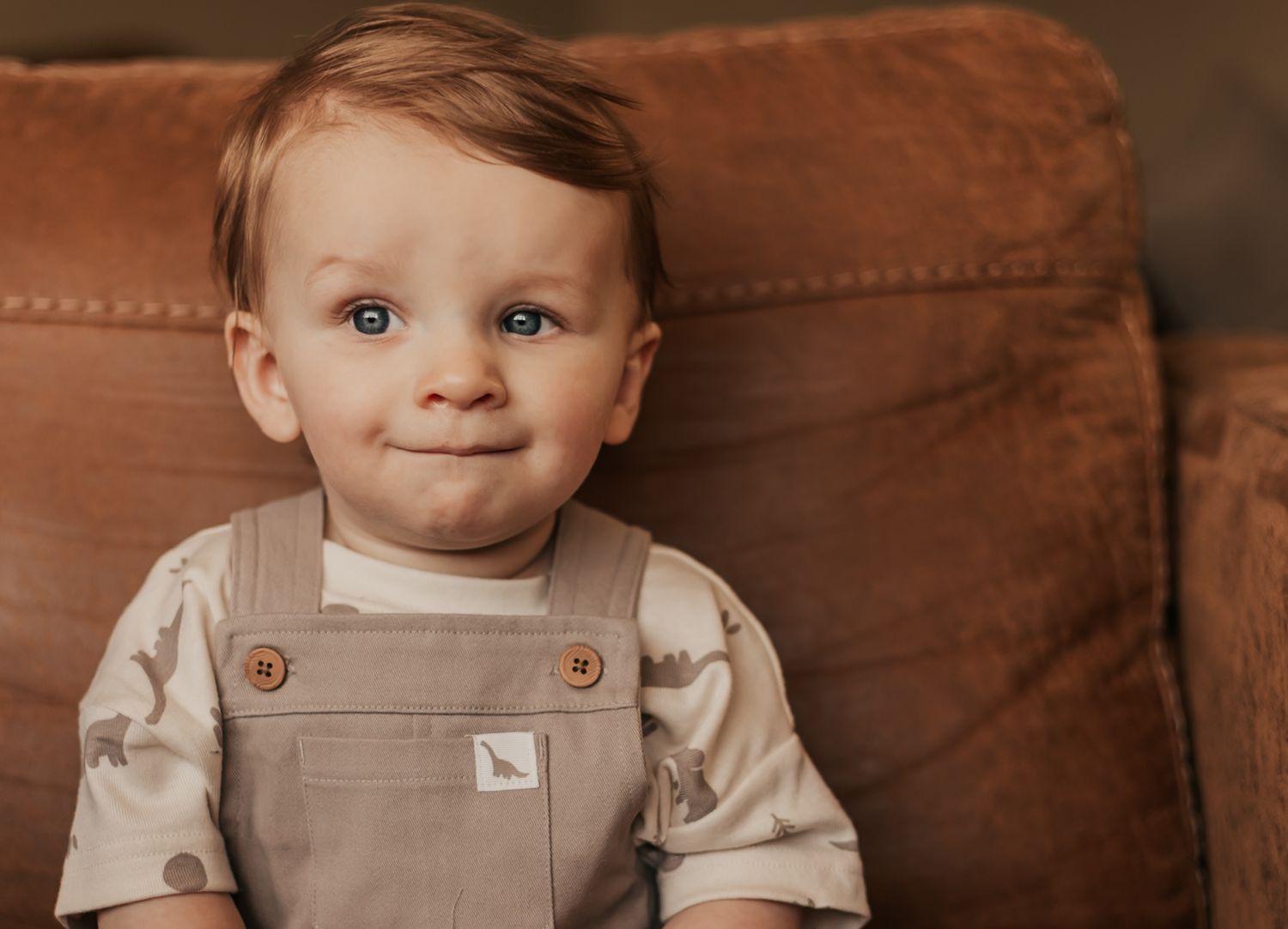 Smiling toddler boy with blue eyes wearing beige overalls sits in front of a brown leather couch | Professional Milestone Sessions for Baby