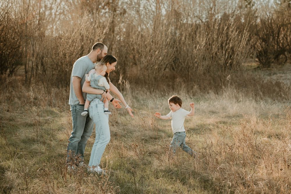 Family of four enjoying a golden hour outdoor photo session in a field, parents holding baby while toddler runs toward them.