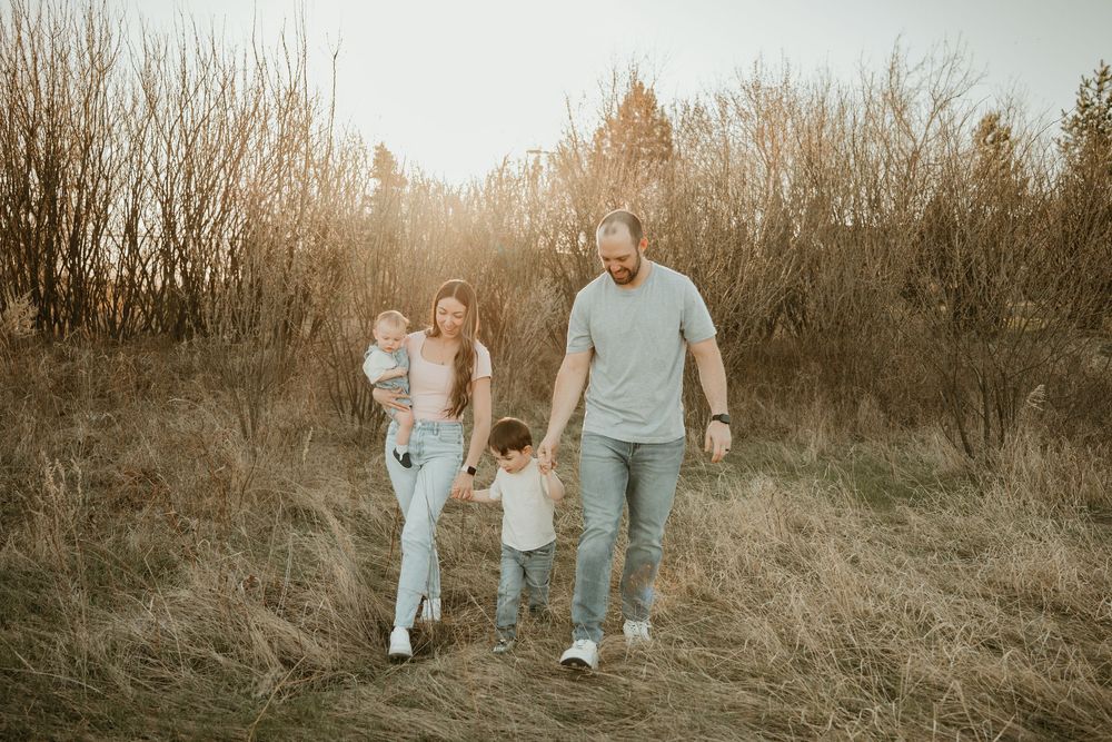 Family of four walking through golden field at sunset, parents holding children's hands among tall grass and bare trees.