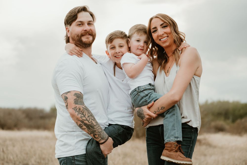A happy family of four poses outdoors in a field, parents with tattoos holding two young boys, wearing white shirts.