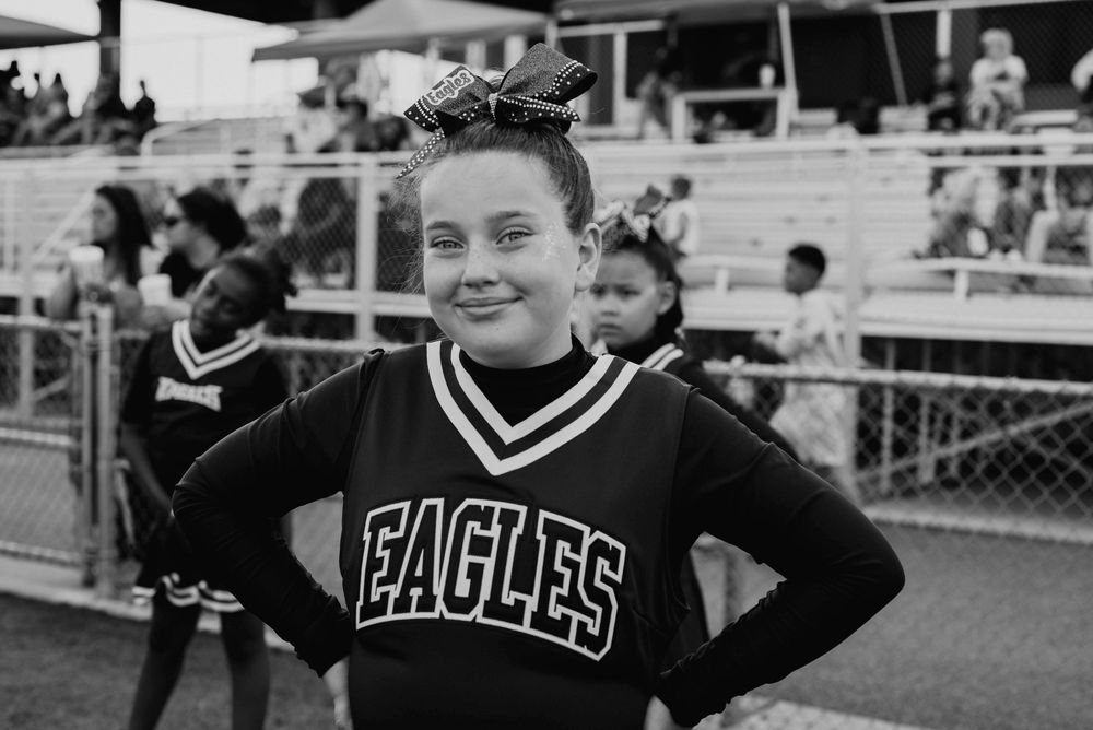 Young girl in Eagles cheerleader uniform smiling with hands on hips at football game.