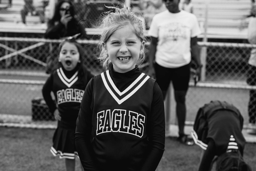 Young girl in Eagles cheerleader uniform smiling brightly on field with teammates in black and white photo.