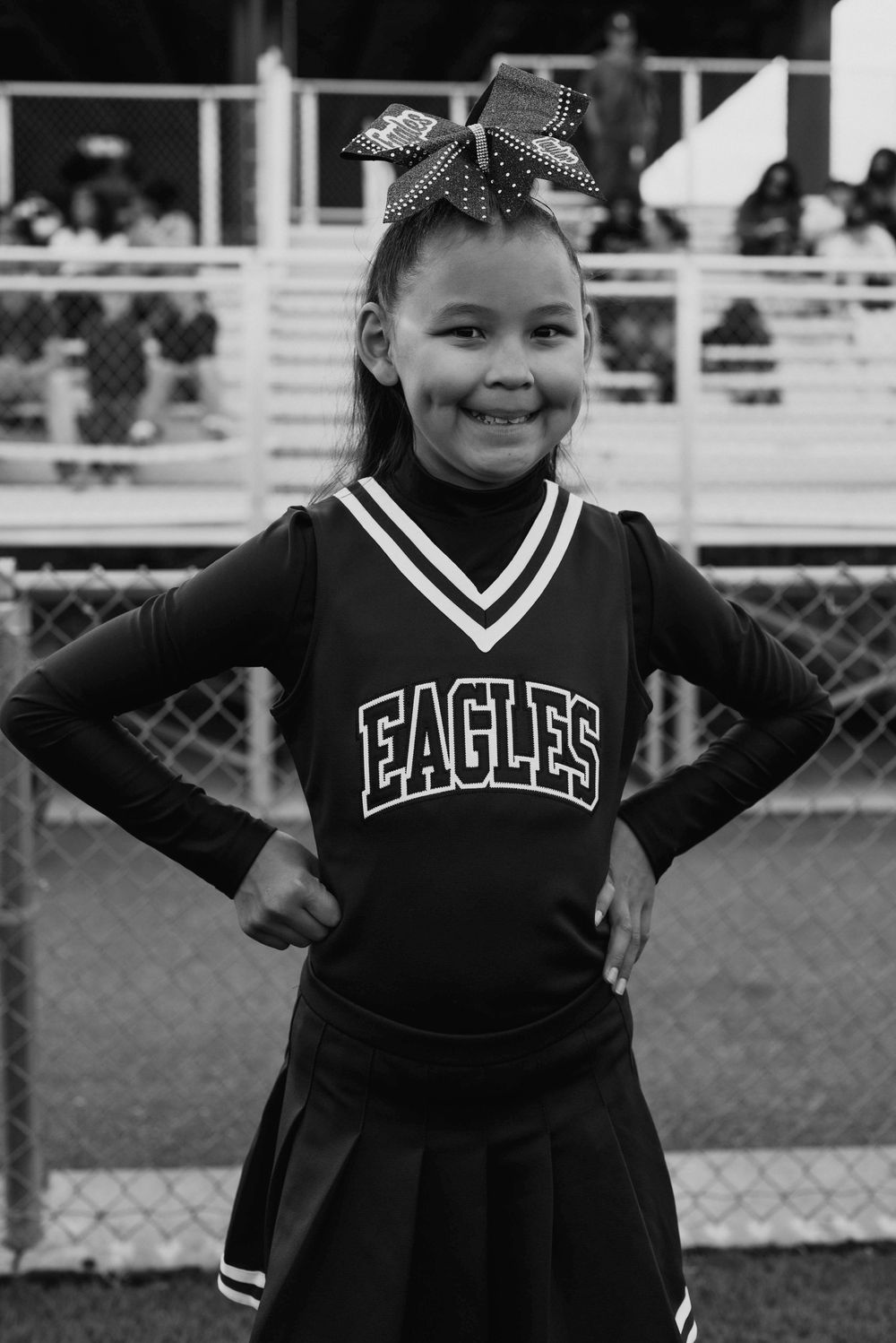 Young girl in Eagles cheerleader uniform with bow smiles proudly at stadium, black and white photo.