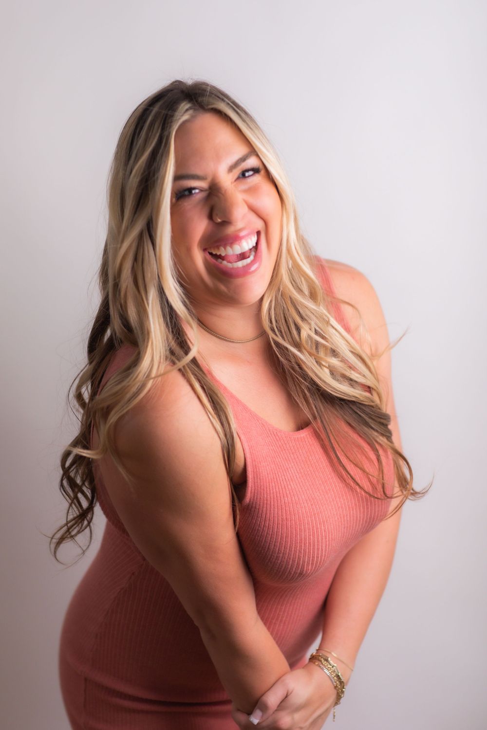 Smiling blonde woman in a pink ribbed dress posing confidently against a light gray studio background.