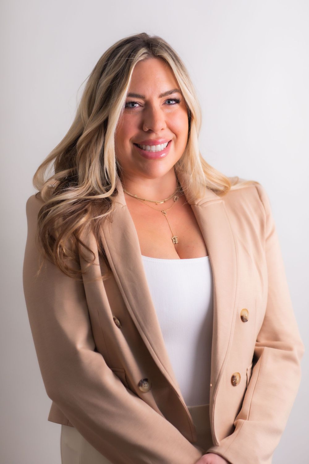 Smiling blonde woman in beige blazer and white top with gold necklace poses for professional headshot.