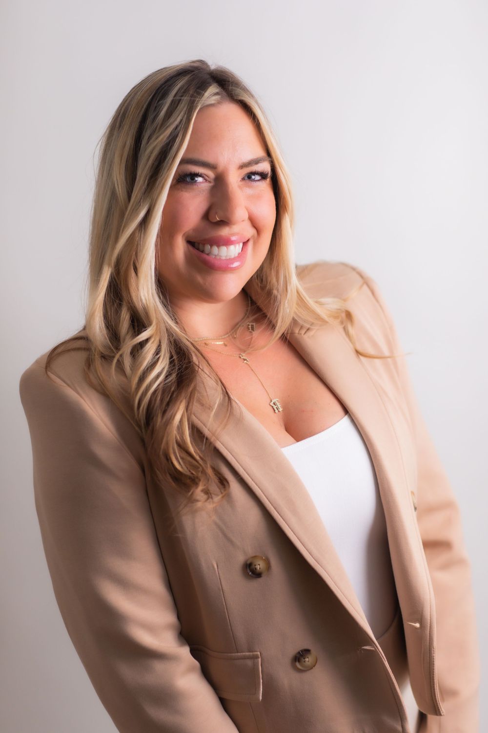 Smiling blonde woman in beige blazer and white top posing against light gray background.