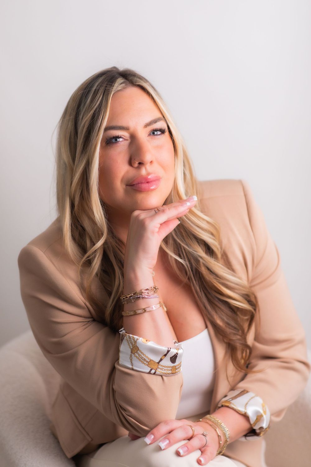 Blonde woman in beige blazer with gold bracelets poses elegantly blowing a kiss in professional photo.