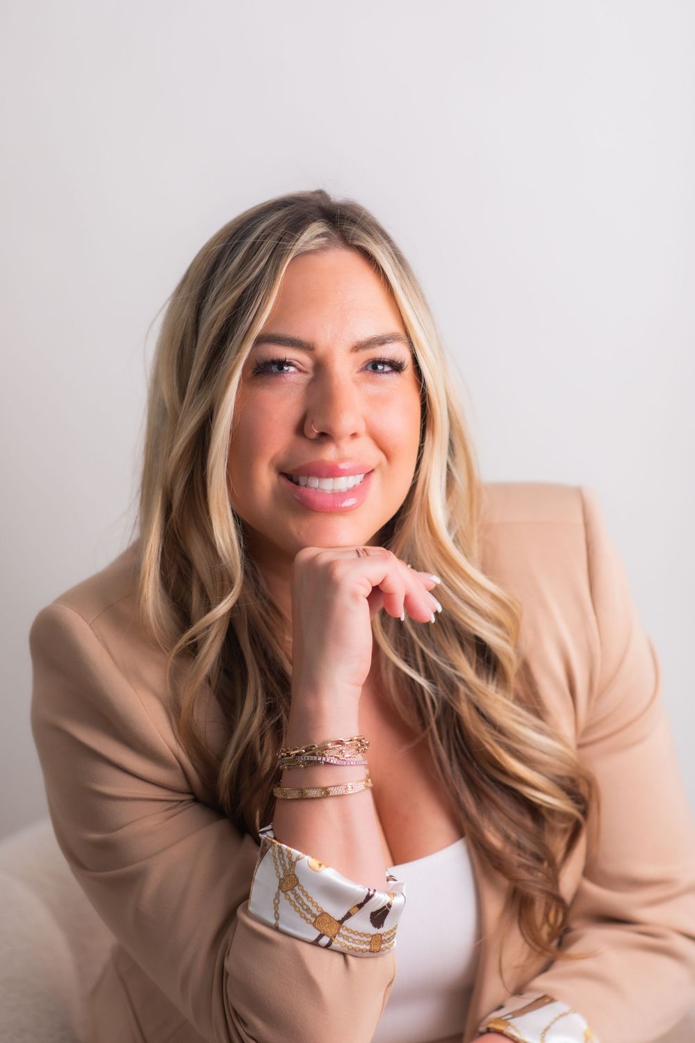 Professional blonde woman in beige blazer smiling confidently with hands clasped in a bright studio setting.