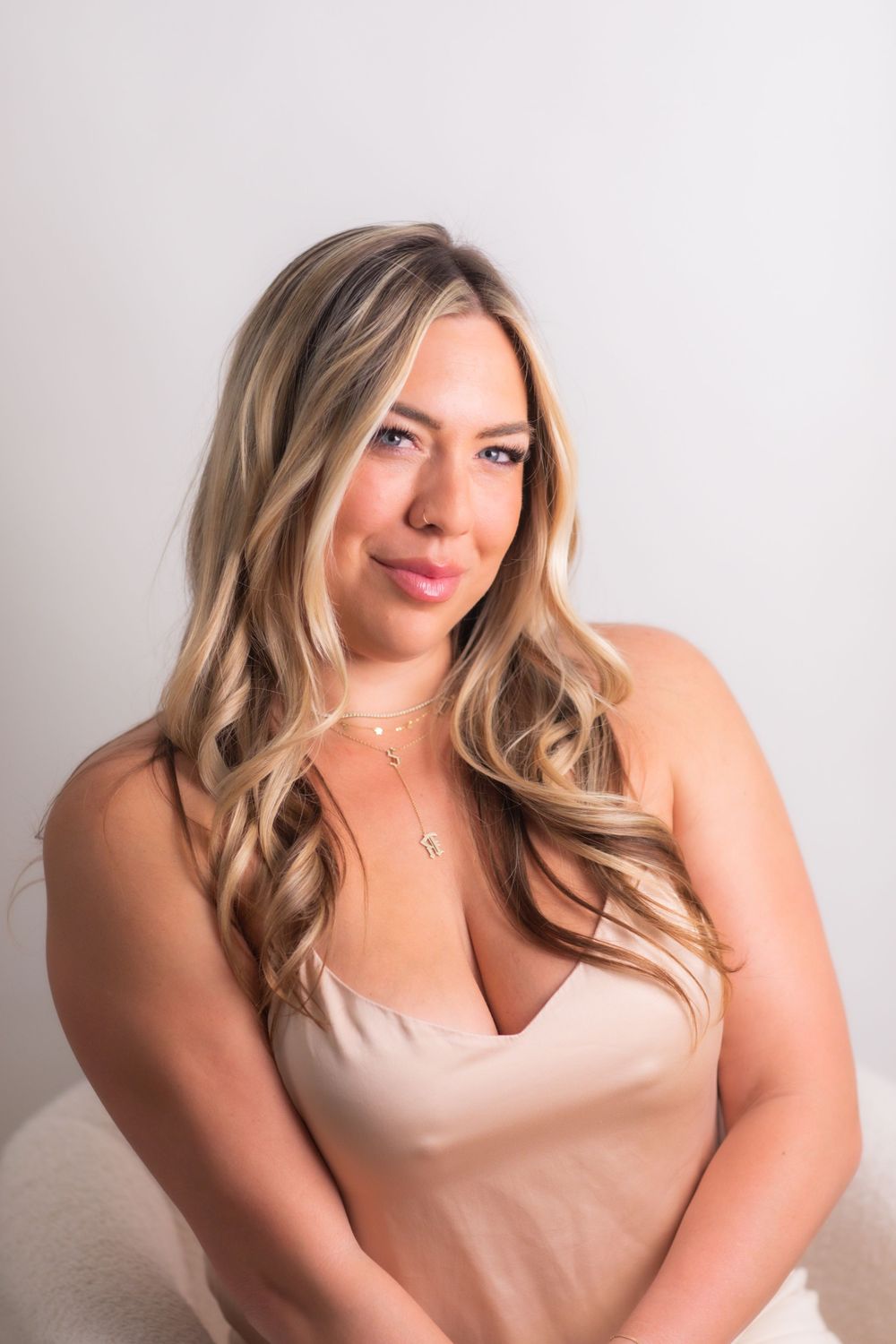 Blonde woman in beige satin top with layered necklaces posing confidently against white background.