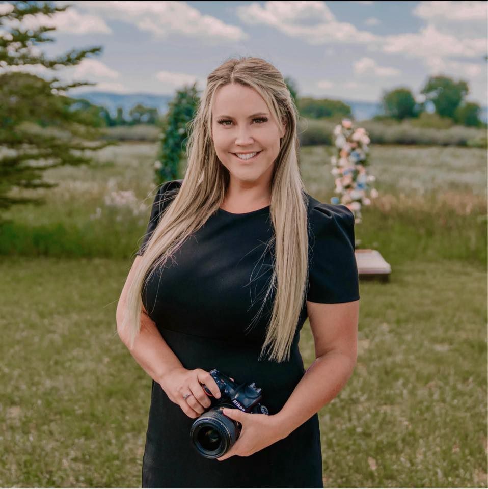 Smiling blonde woman in black dress holding a camera outdoors in a green field at a wedding venue.