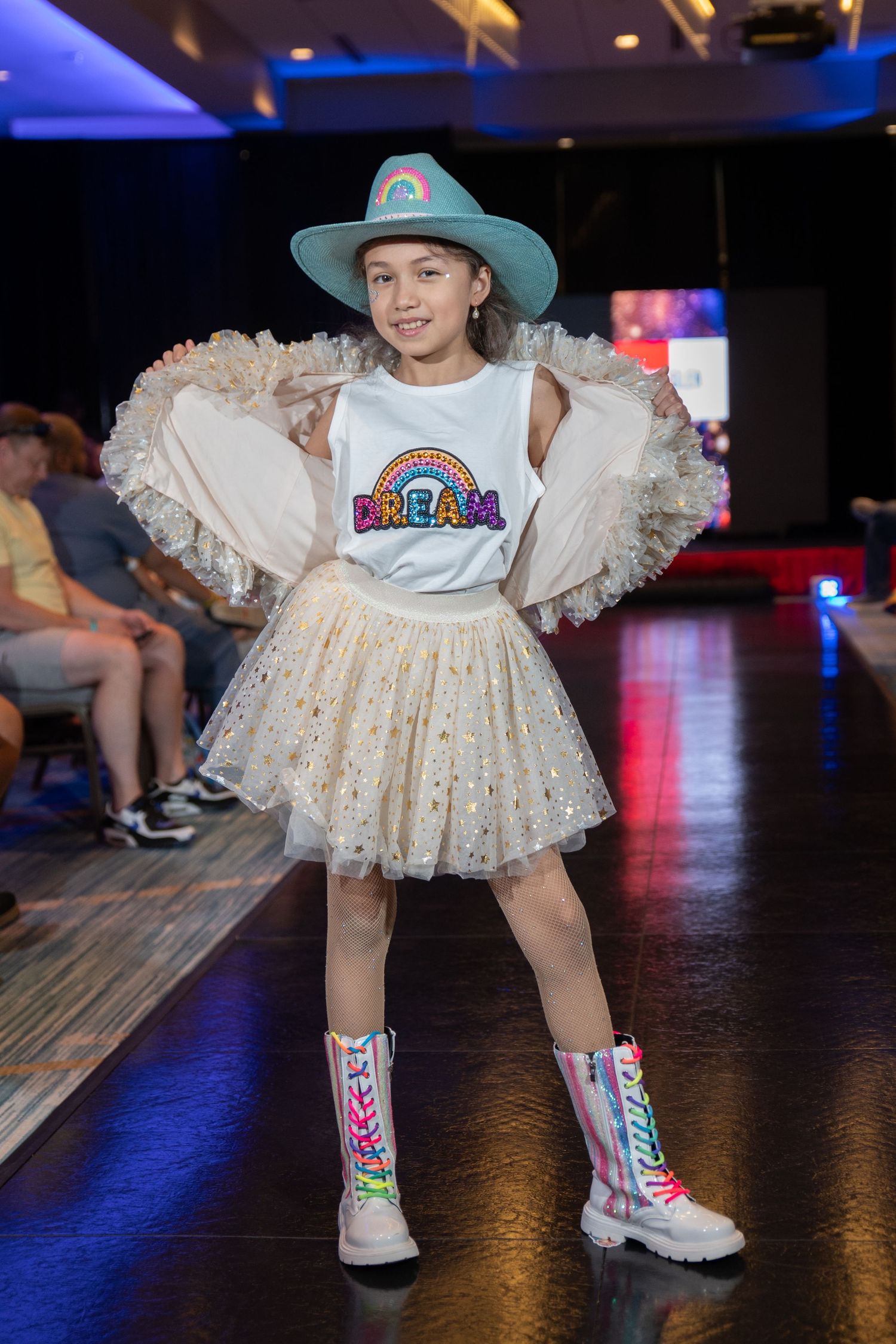 Young girl modeling a DREAM graphic tee, tutu skirt, teal cowboy hat, and colorful boots on a runway.