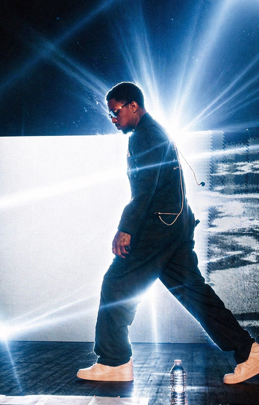 Performer in dark jumpsuit and sunglasses walks on stage with dramatic bright light beams behind them.
