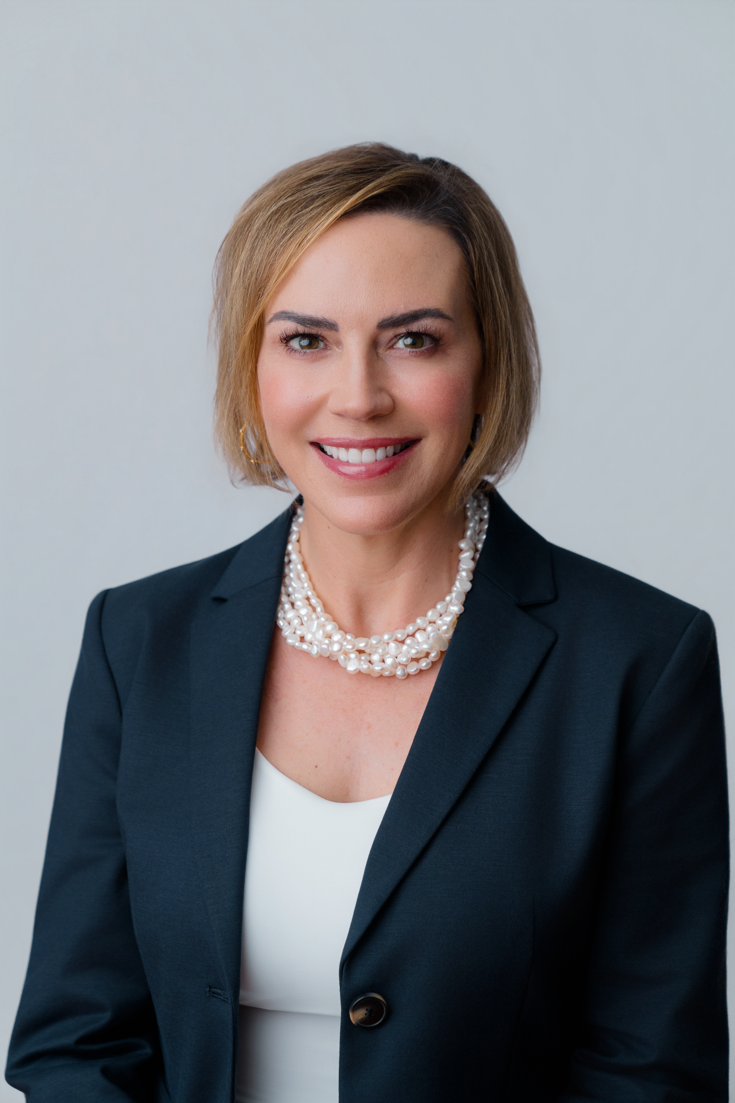 Professional woman in dark blazer and pearl necklace smiling against gray background.