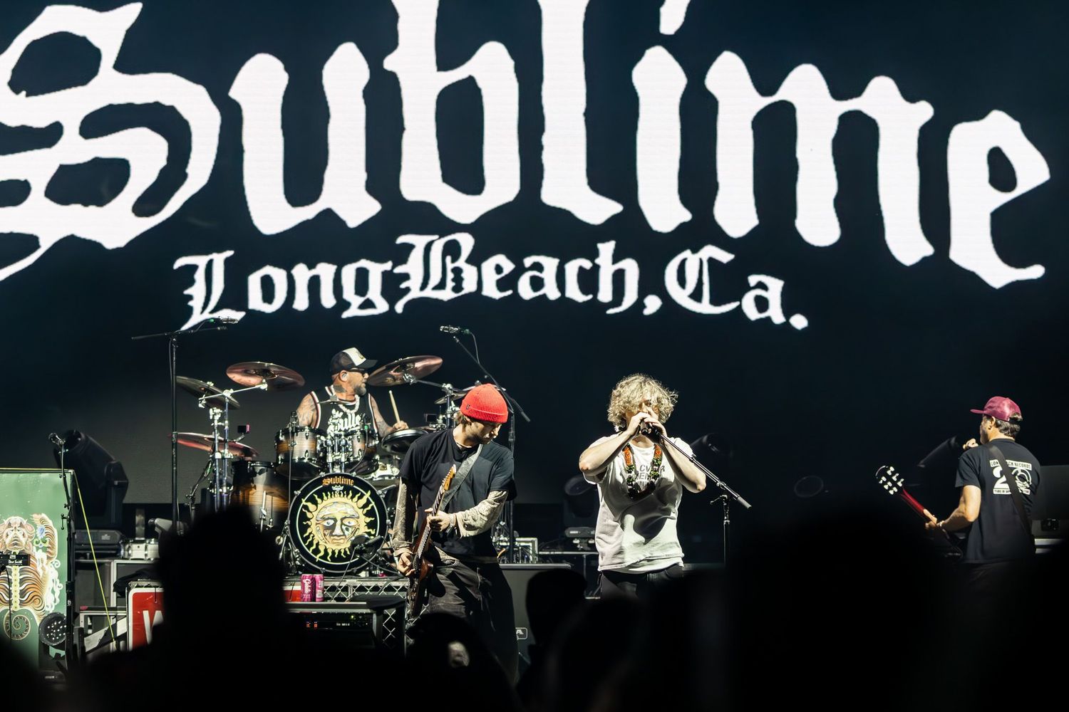 Live music photography of the band Sublime performing on stage in front of a giant "Sublime Long Beach, Ca." backdrop.