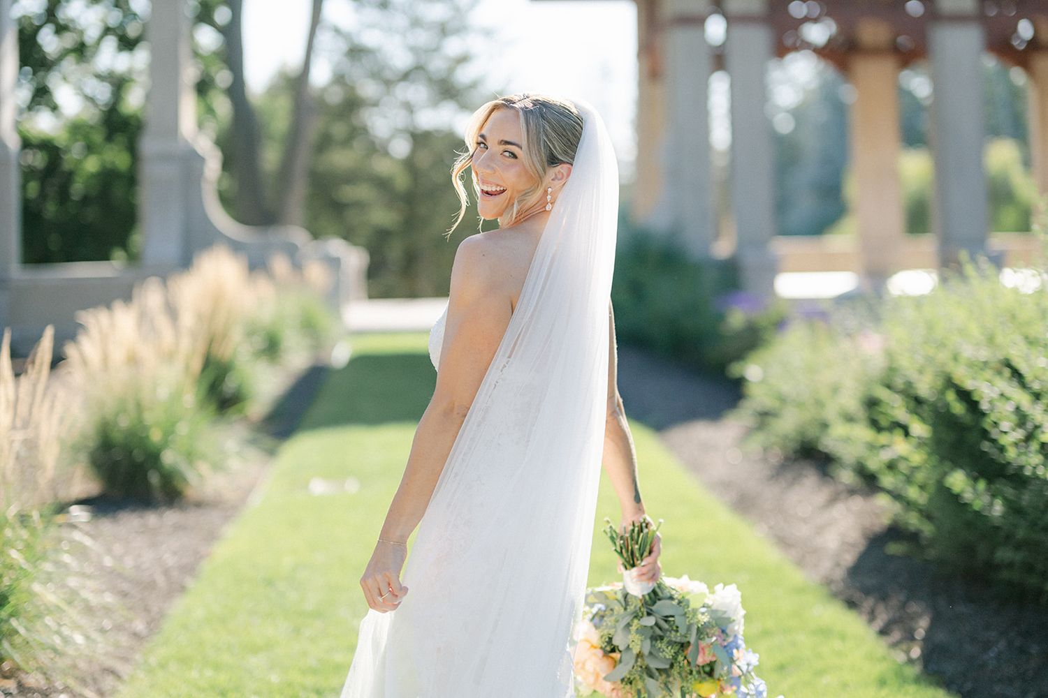 Armour House Wedding - Bride in lace mermaid gown walks garden path holding bouquet, elegant columns and greenery backdrop.