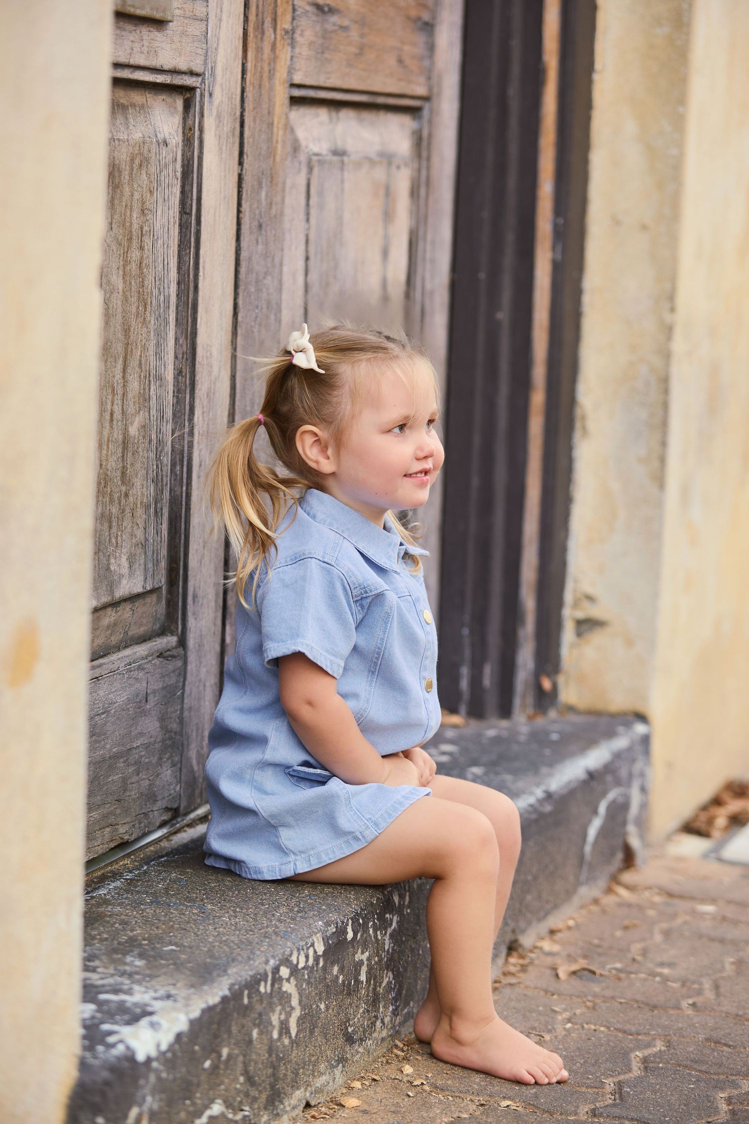 Young barefoot girl in blue dress sitting on stone steps beside a rustic wooden door.