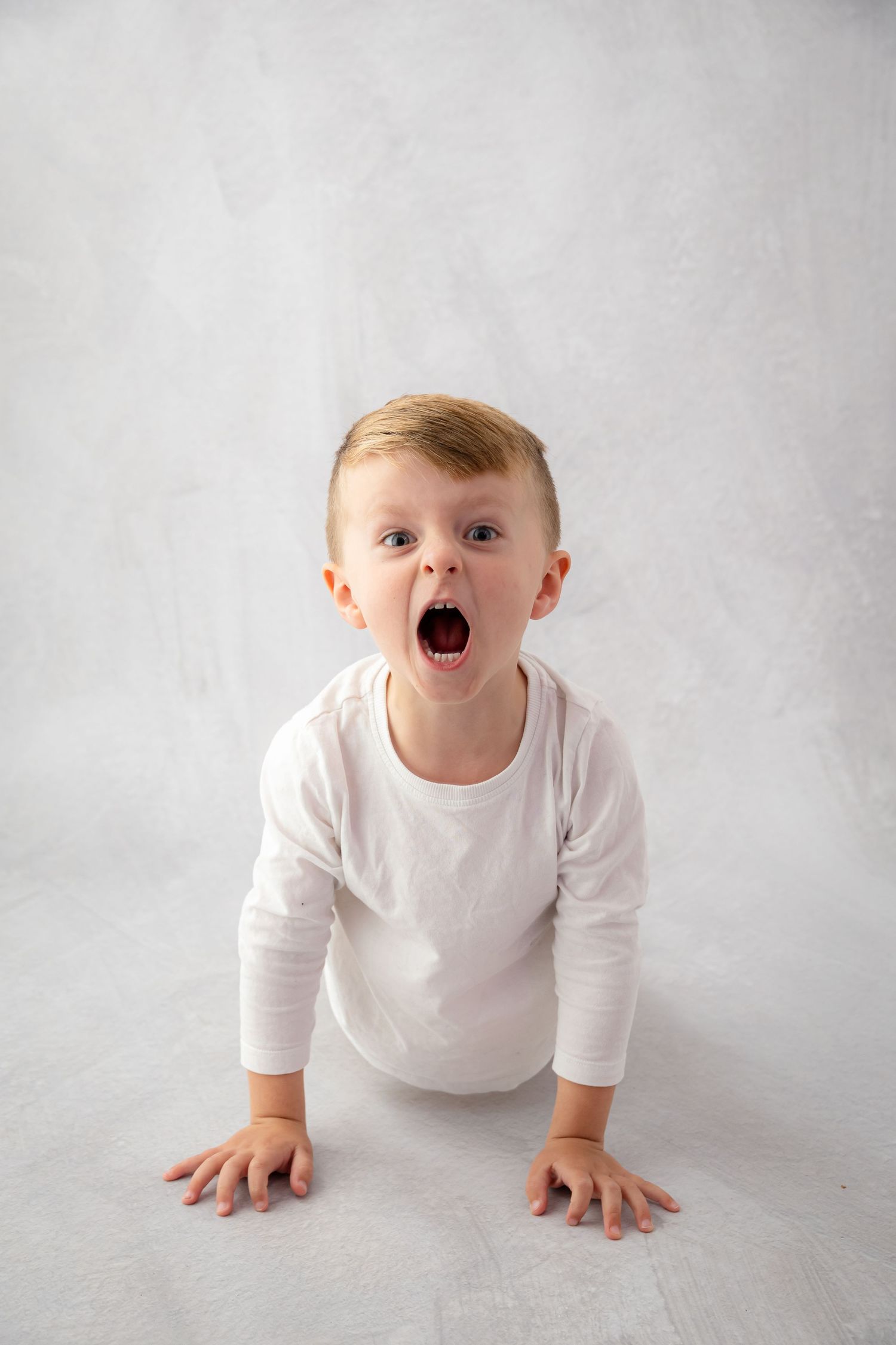 Surprised toddler boy in white long-sleeve bodysuit kneeling on floor with mouth wide open against gray background.