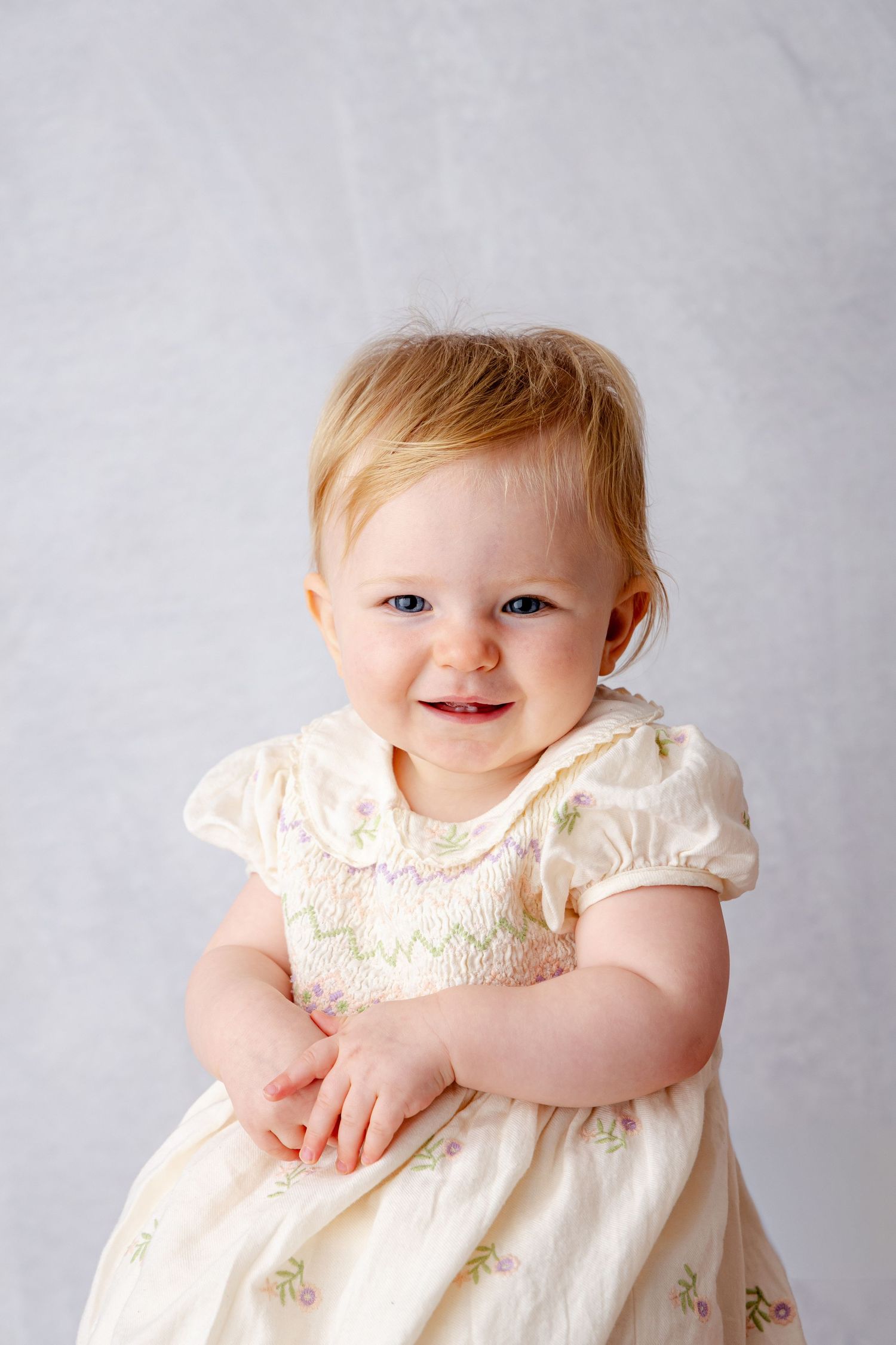 Smiling baby girl with blue eyes wearing a cream floral smocked dress, seated against a light gray background.