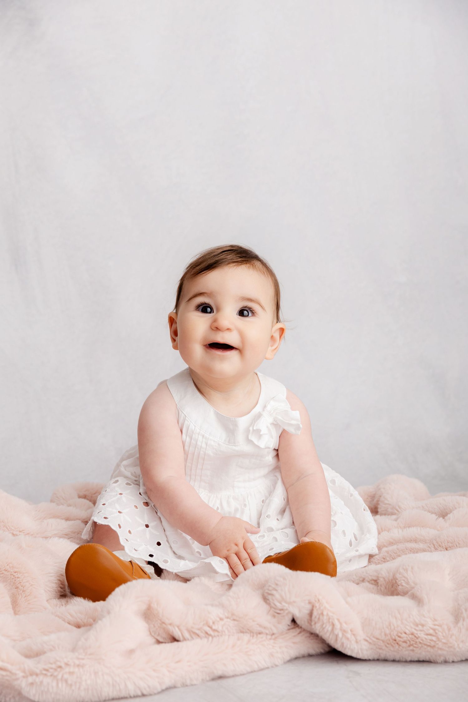 An adorable baby girl in a white dress and brown shoes sits smiling on a pink fluffy blanket.