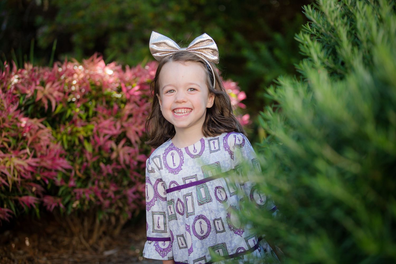 Smiling girl with gold bow headband wearing printed dress, posing outdoors among pink flowers and green shrubs.