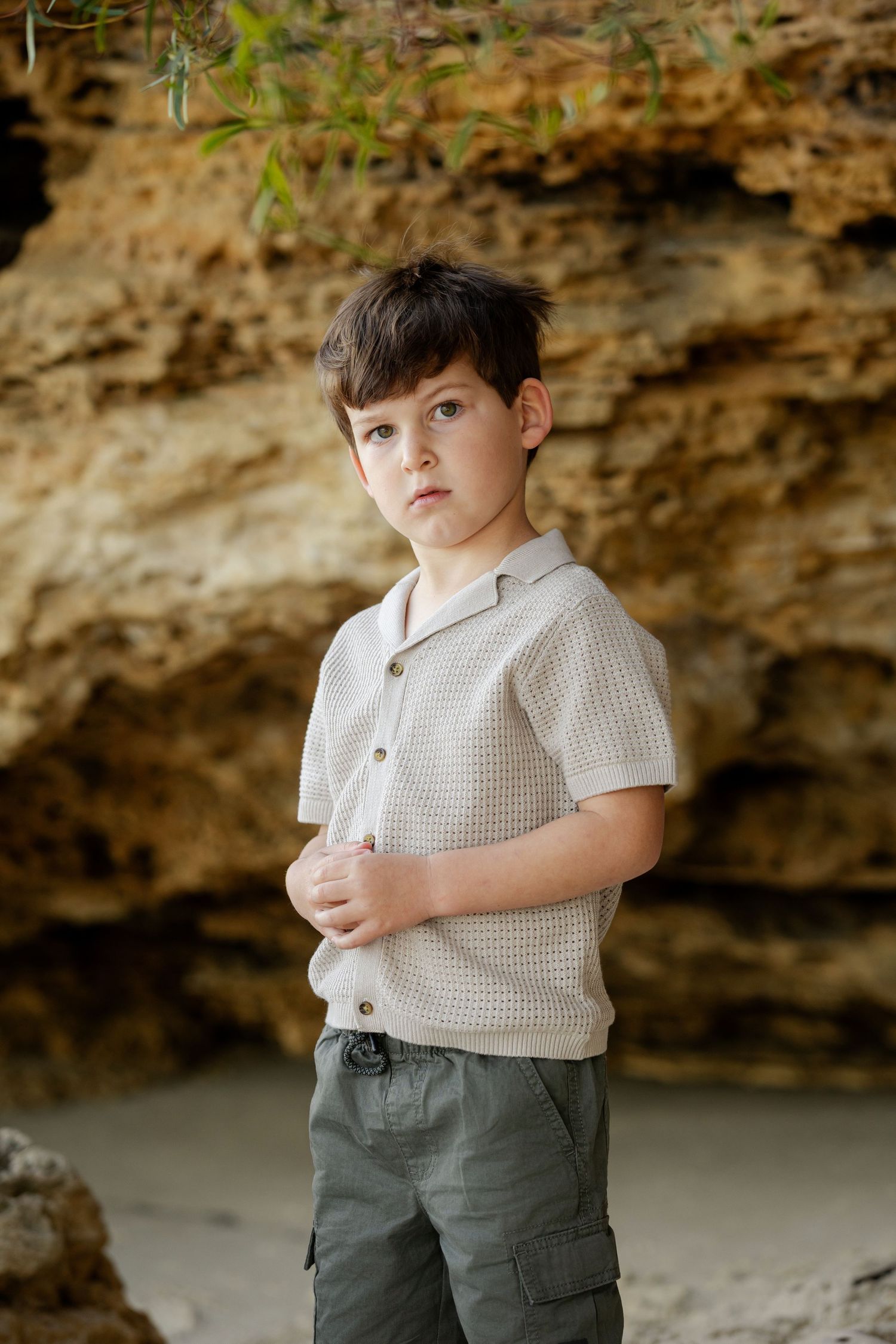 Young boy in beige knit shirt and cargo pants poses against sandy rock formation outdoors.