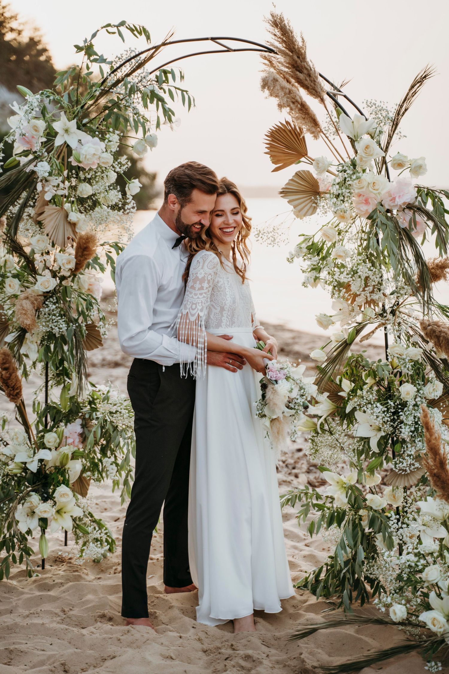 Elegant couple standing under a floral arch on a sandy beach at sunset for a boho wedding ceremony.