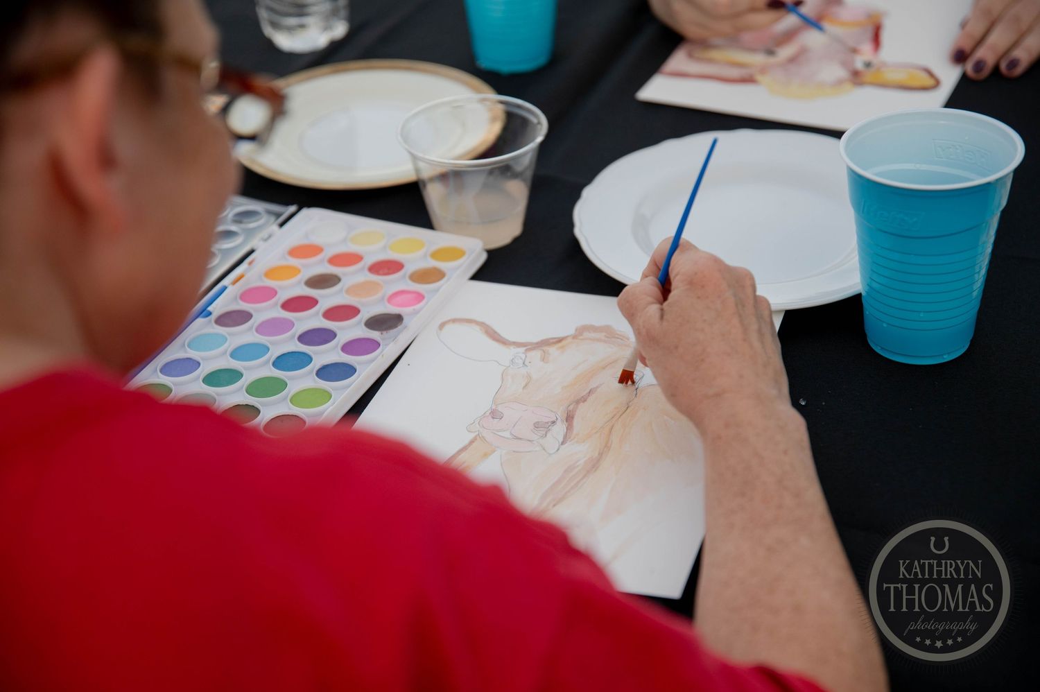 Group of people painting at an outdoor long table event with blue cups and art supplies in a field.