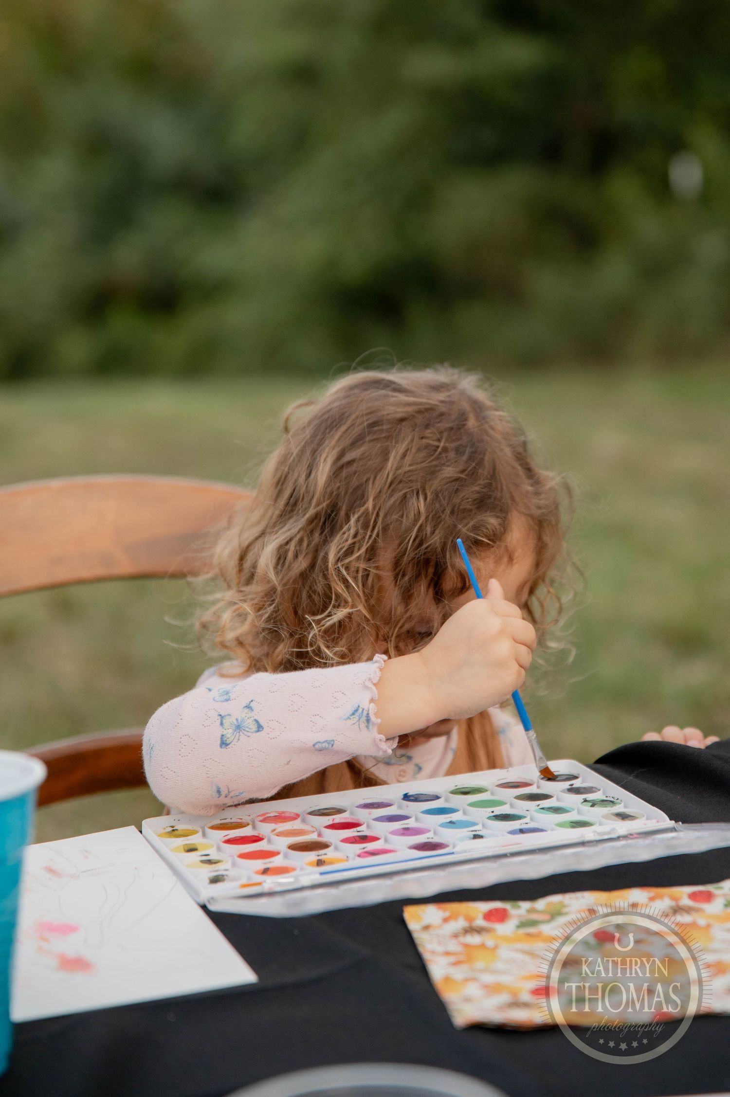 Young child with curly hair painting with watercolors outdoors at a black table, focused on her colorful art project.