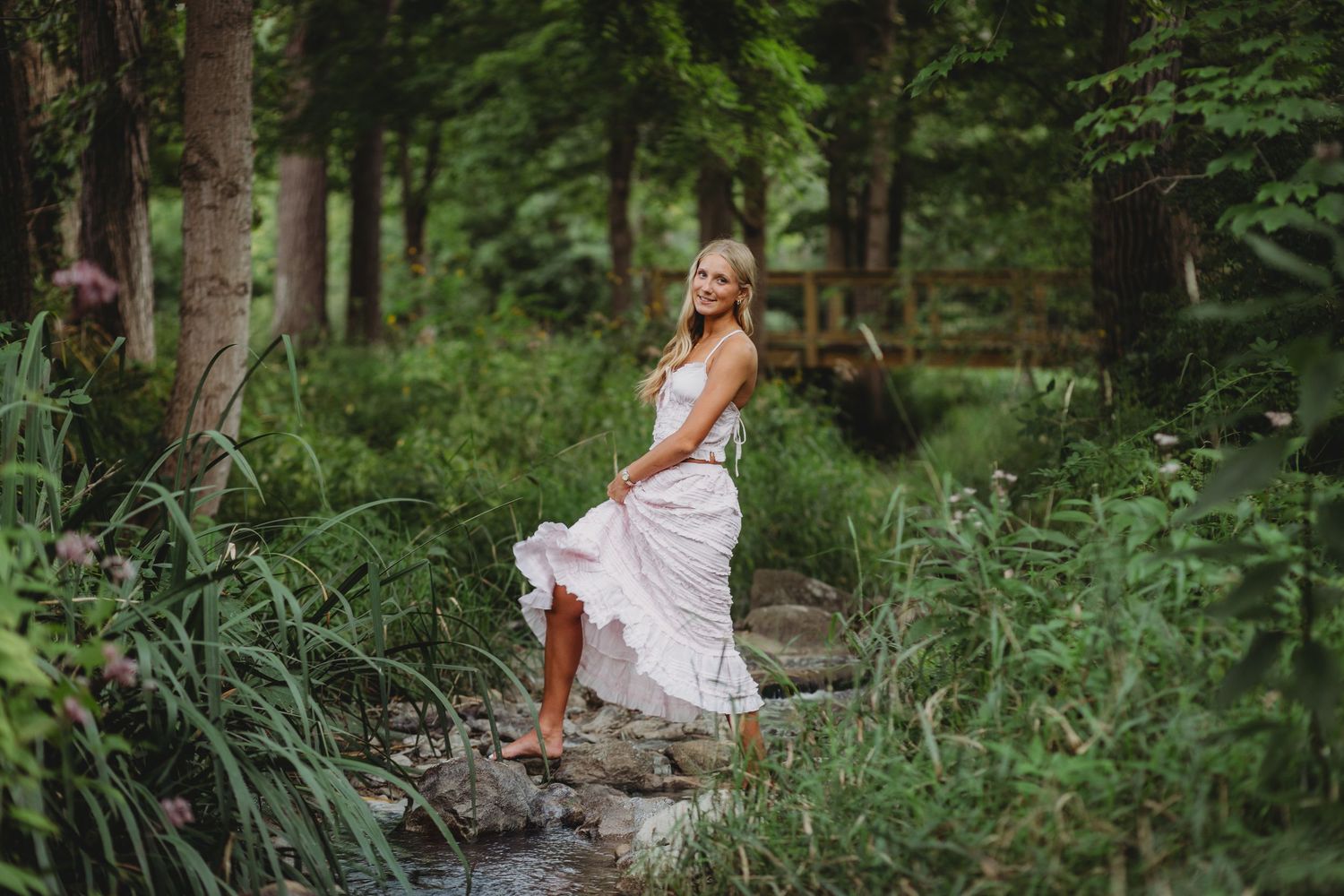 Blonde woman in white dress standing on rocks by a stream in a lush green forest setting. Rochester senior session