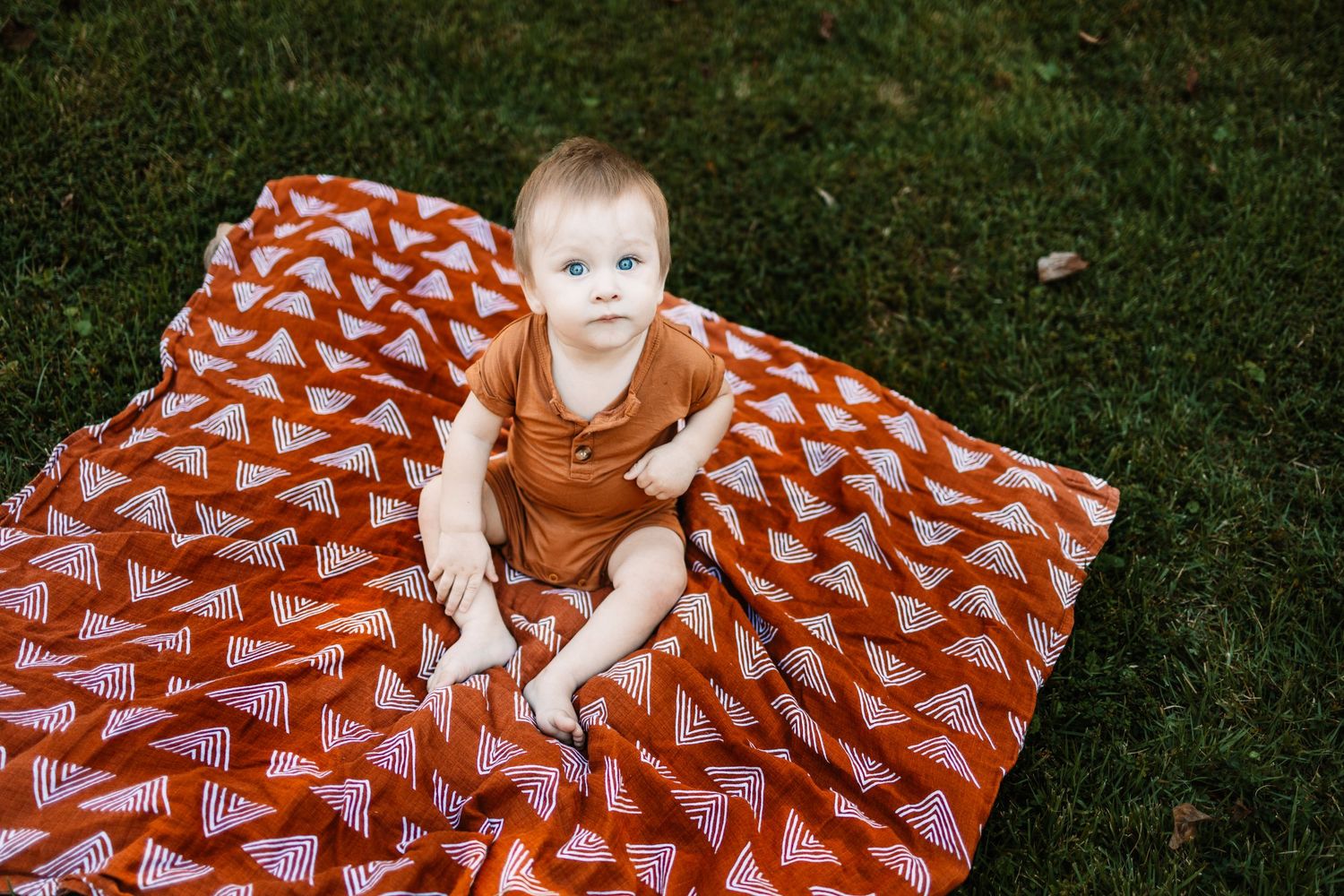 Baby sitting on a rust orange triangle print blanket on green grass outdoors.