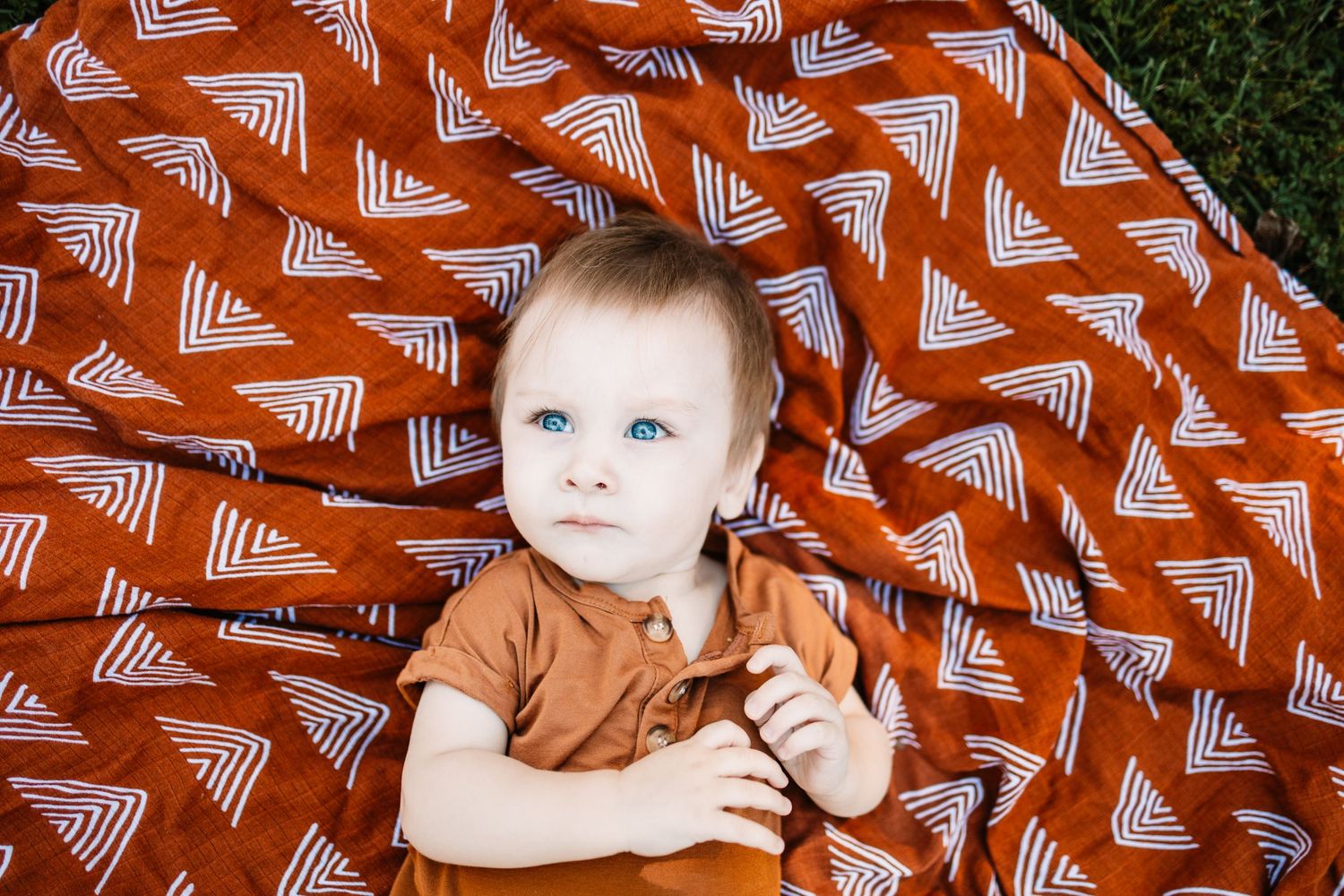 Blue-eyed baby lying on a rust orange patterned blanket with white triangle designs, wearing an orange outfit.