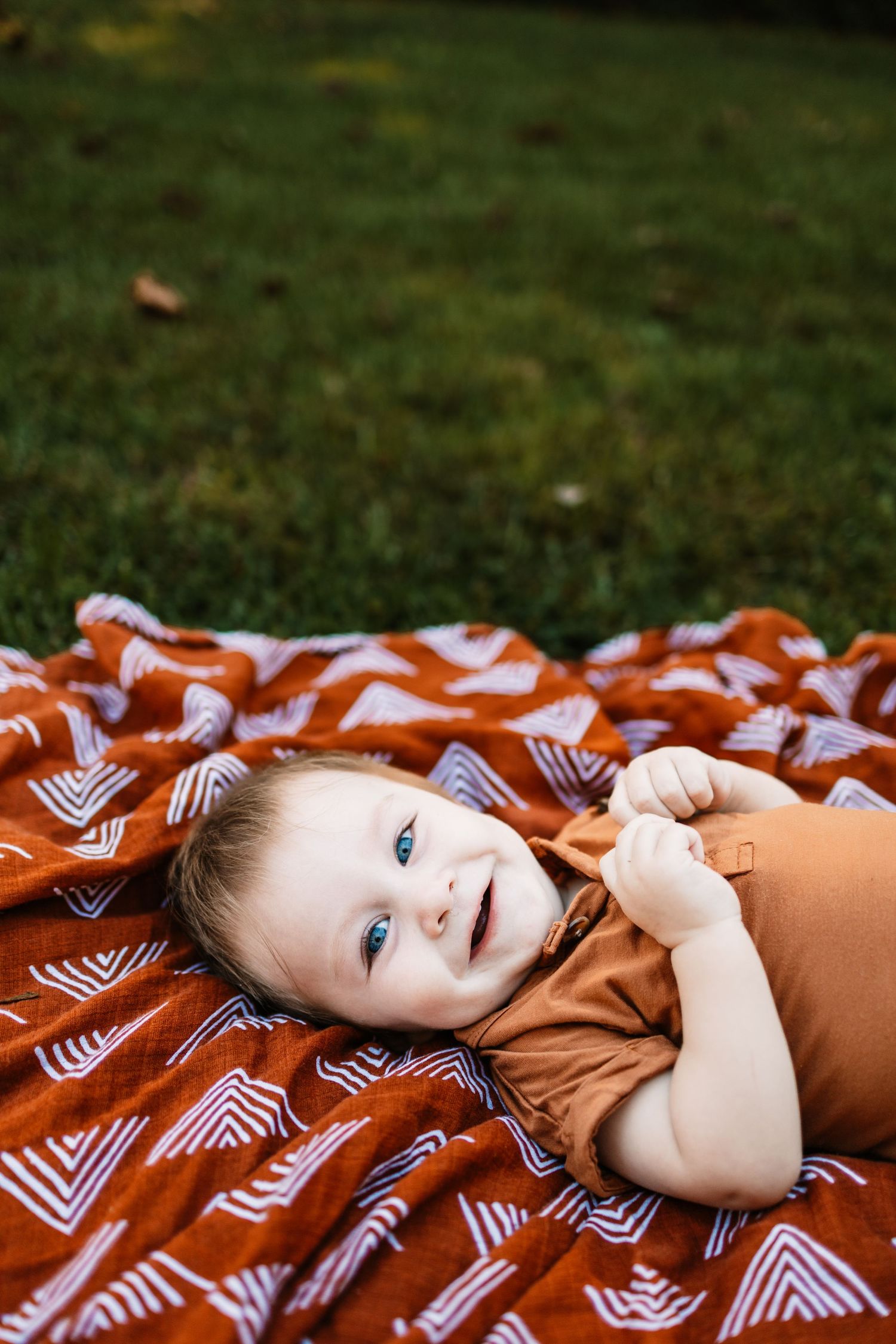 Smiling baby with blue eyes lying on a rust orange patterned blanket on green grass outdoors.