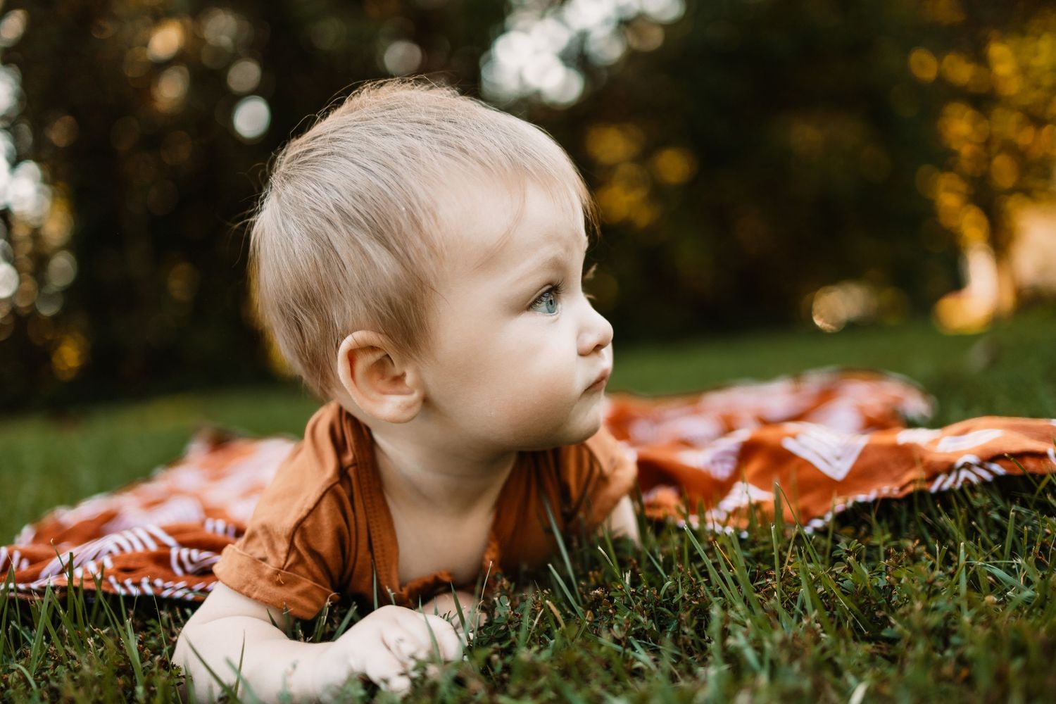 A blonde toddler lies on a red blanket in green grass, gazing curiously in a sunny outdoor setting.