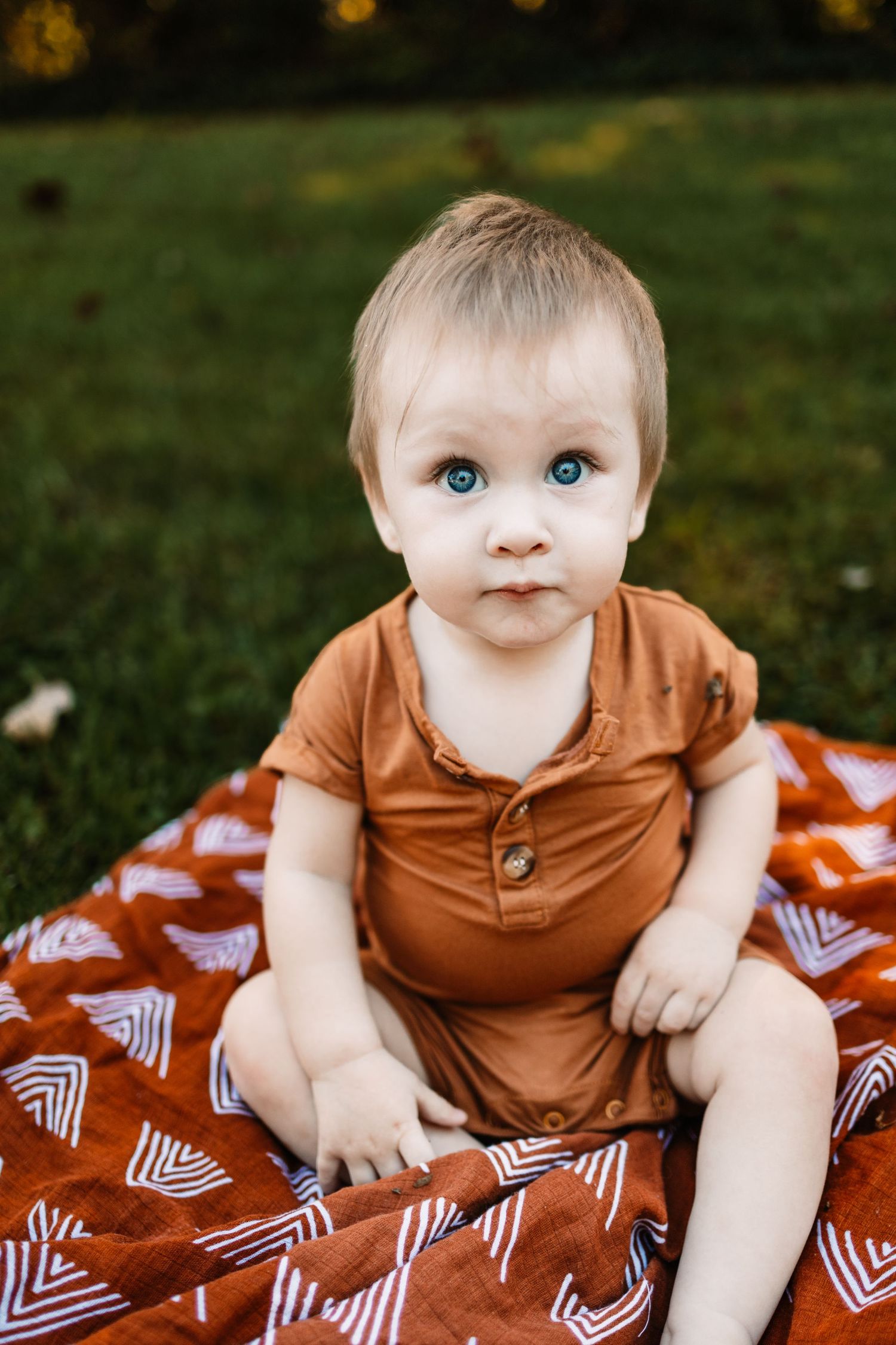 Adorable blue-eyed baby in a rust brown romper sitting on a red patterned blanket on green grass outdoors.