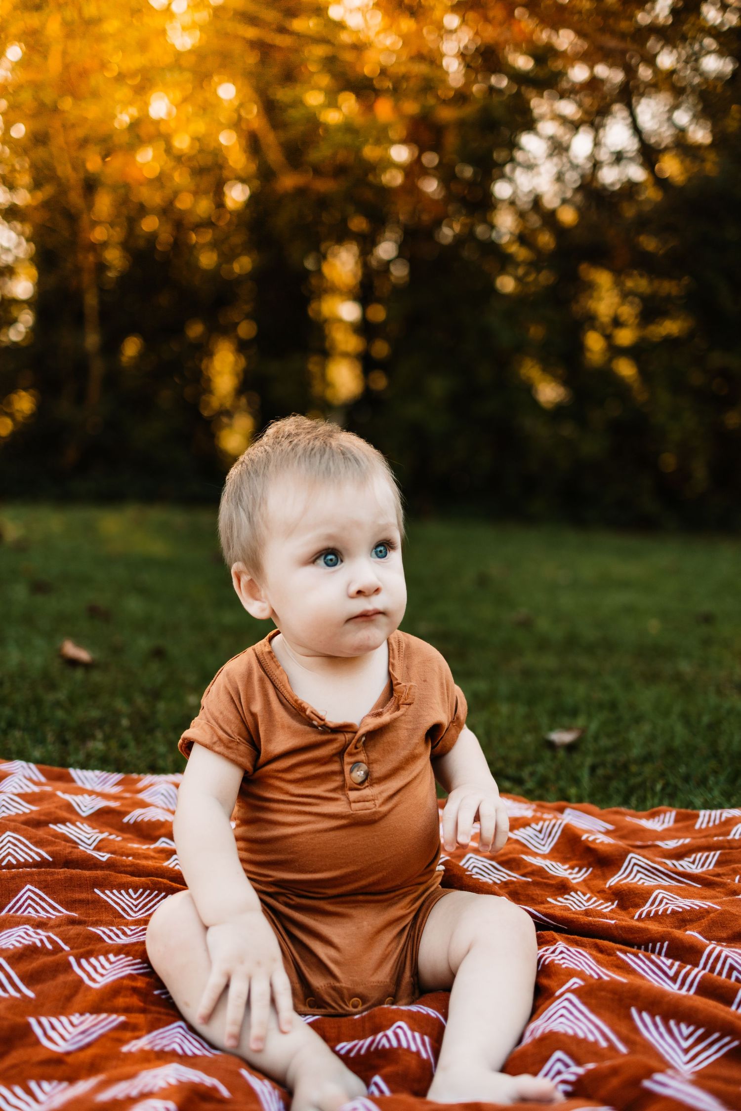 Blonde baby with blue eyes sitting on a red patterned blanket outdoors in autumn sunlight wearing a brown romper.