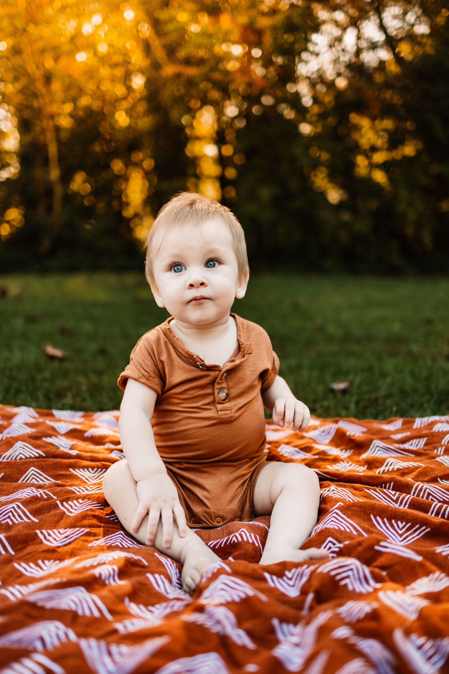 Adorable baby with blue eyes sitting on a red blanket outdoors in golden autumn sunlight wearing a brown romper.