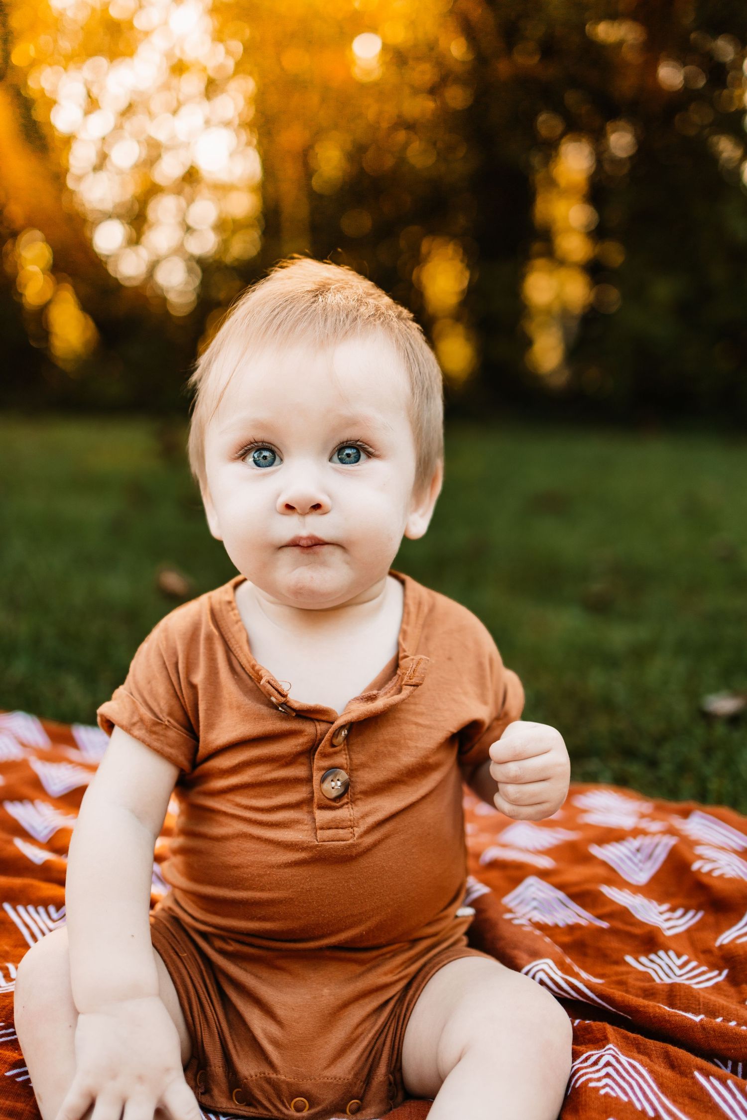 A cute baby with blue eyes sits on a red blanket outdoors wearing a brown romper during golden hour.