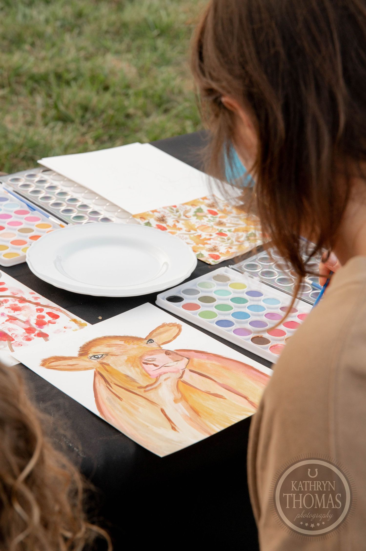 Two people painting watercolors at a black table with blue cups, a charcuterie plate, and art supplies outdoors.