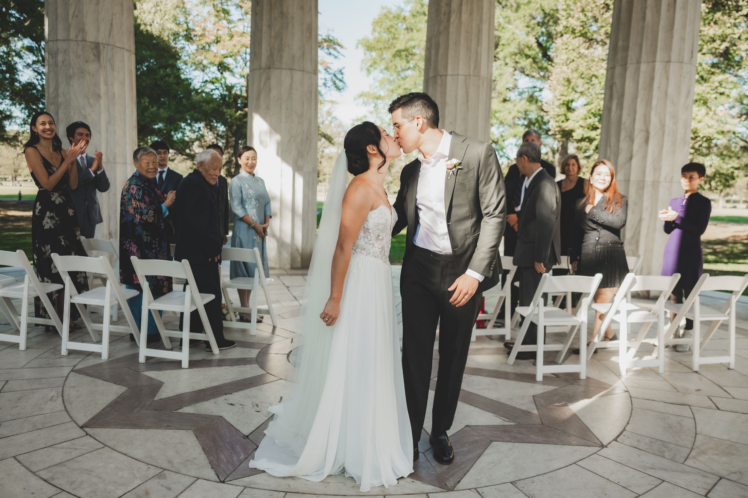 Newlyweds share a first kiss at an outdoor wedding ceremony with white chairs and applauding guests at a columned pavilion.