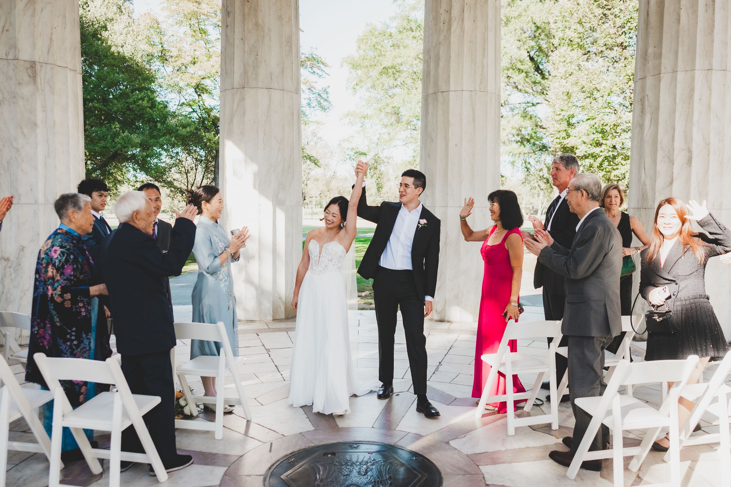 Bride and groom celebrate at outdoor wedding ceremony aisle surrounded by guests at elegant columned venue.