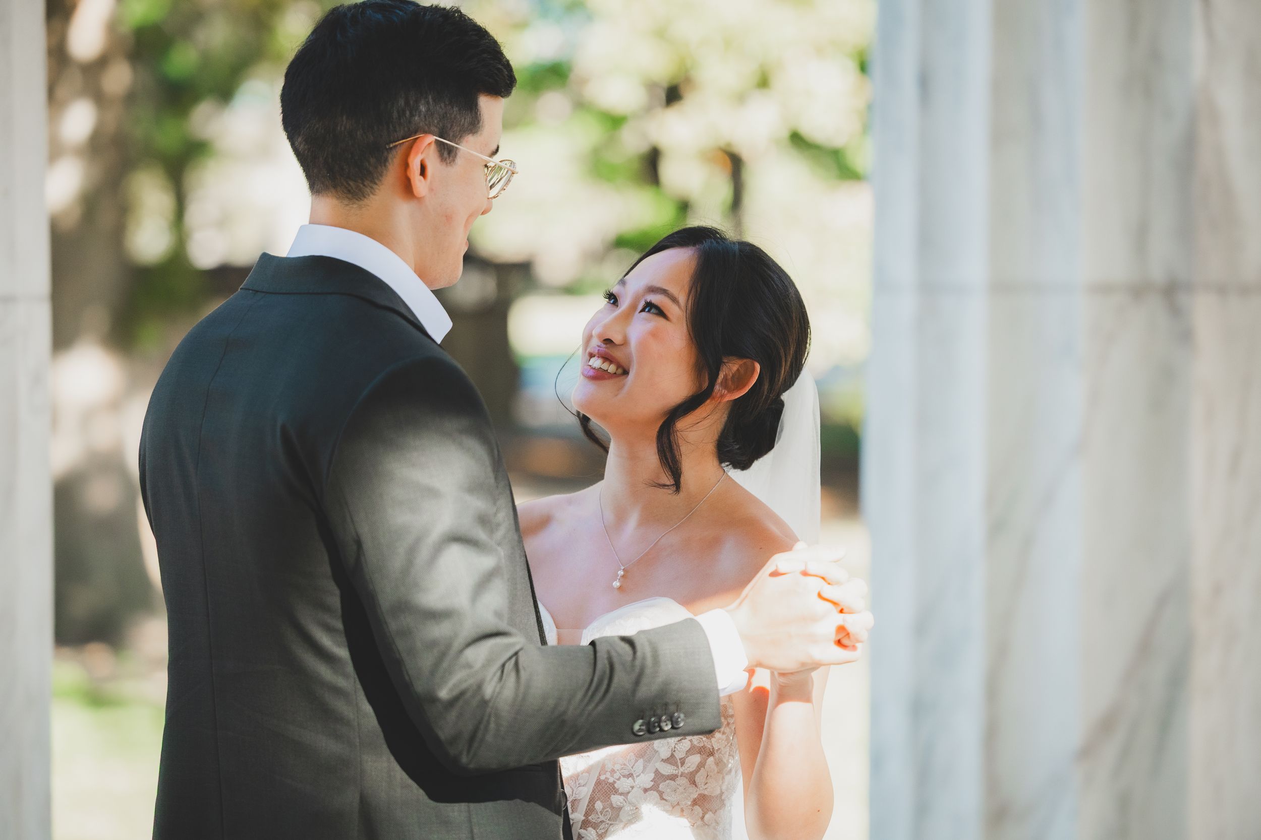 A joyful bride in a white gown smiles at her groom in a dark suit during an outdoor wedding ceremony.