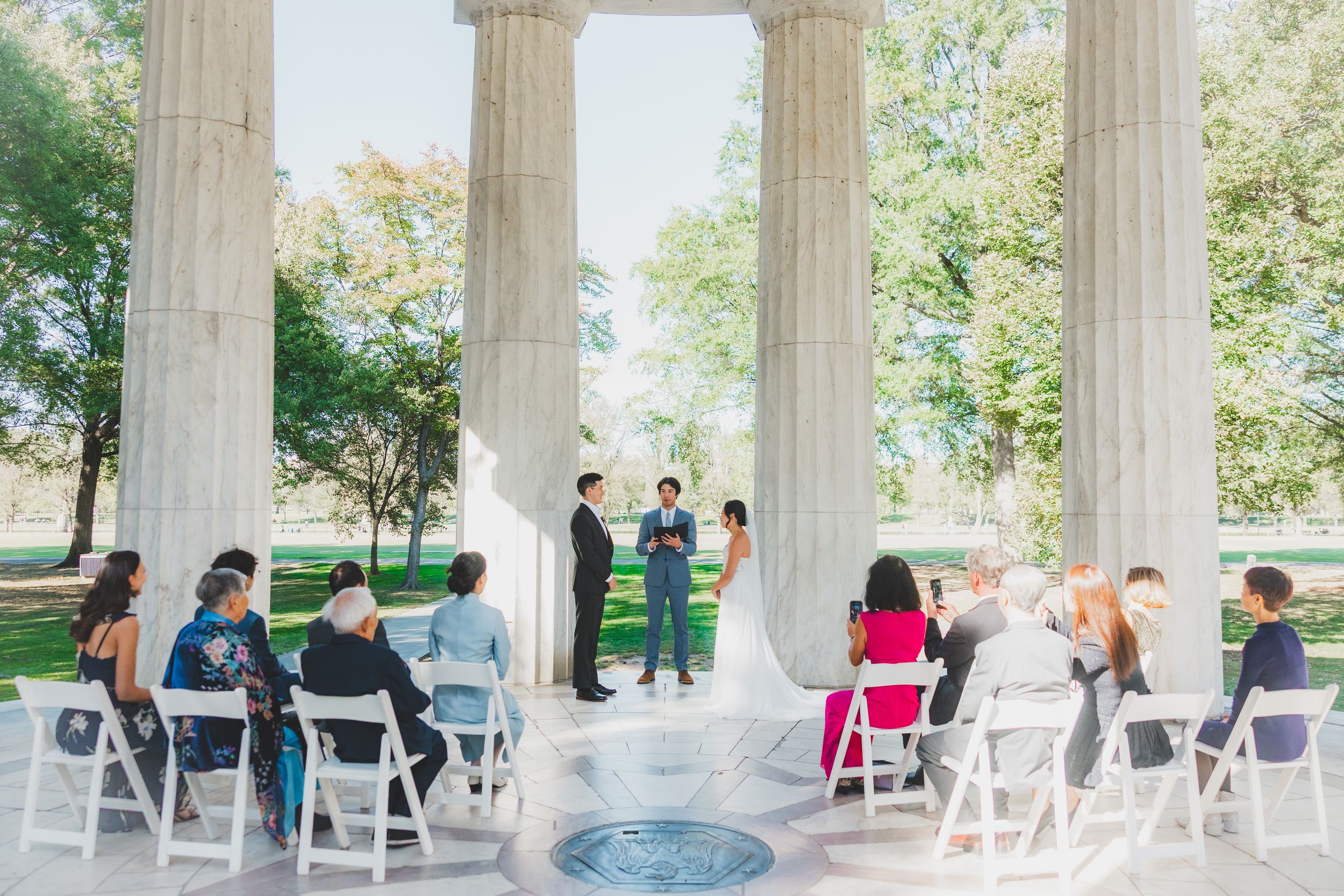 Outdoor wedding ceremony under stone columns with guests seated in white chairs on a sunny day.