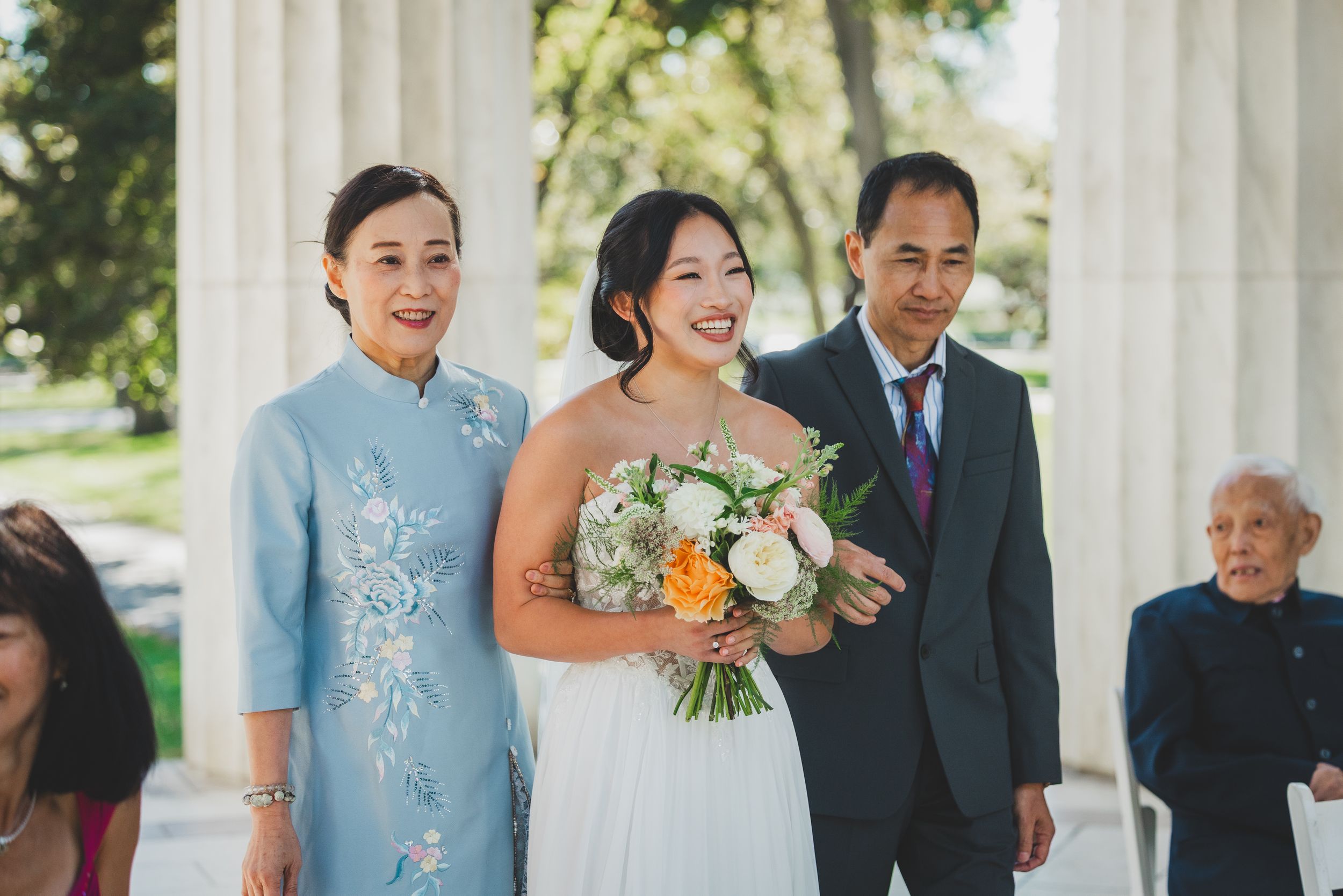 Bride in white gown holds floral bouquet flanked by parents in formal attire at an outdoor wedding ceremony at the dc war memorial
