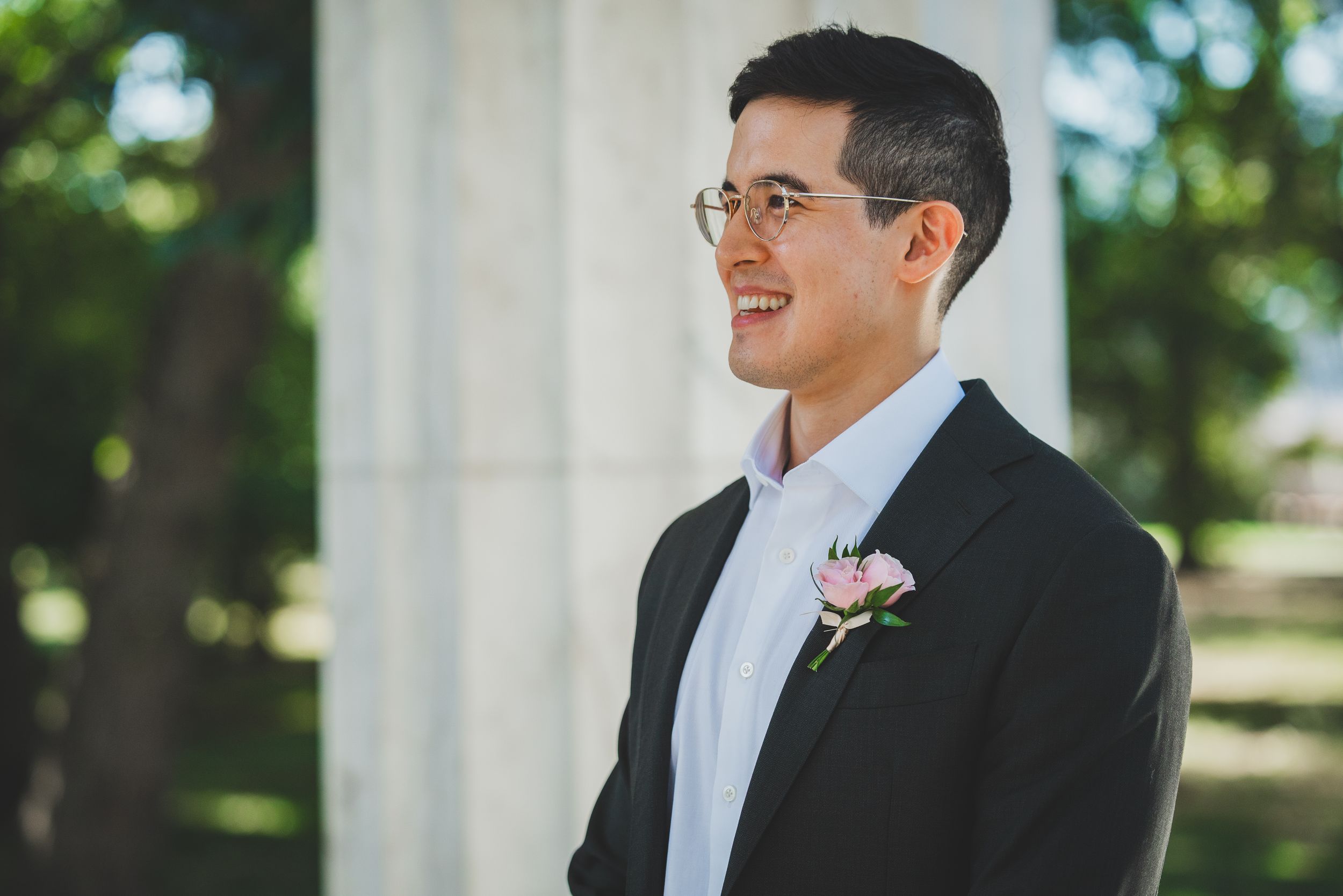 Smiling groom in black suit with pink boutonniere standing outdoors at washington dc wedding at dc war memorial